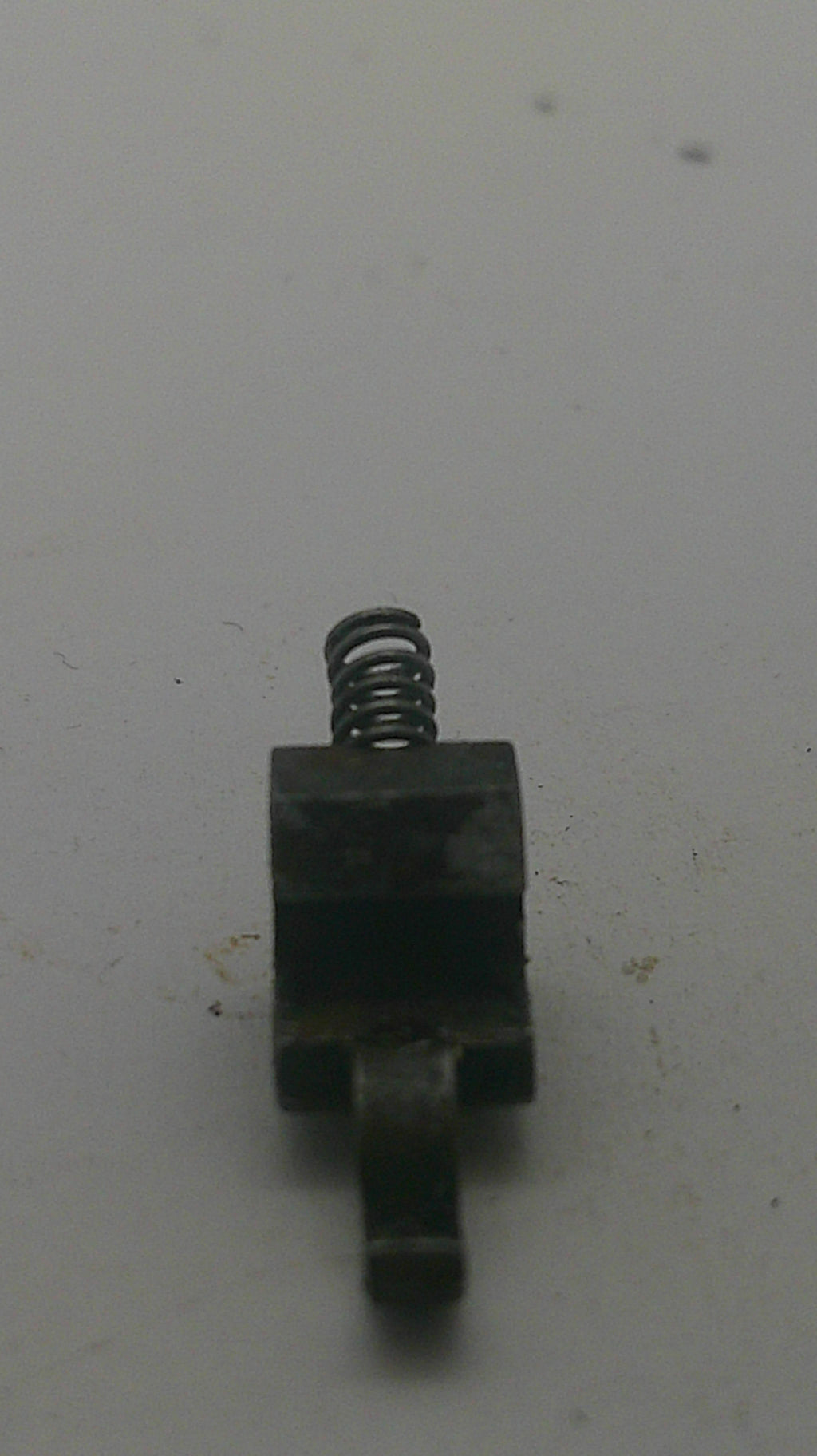A small dark metal mechanical component with a coiled spring, photographed on a plain off-white surface.