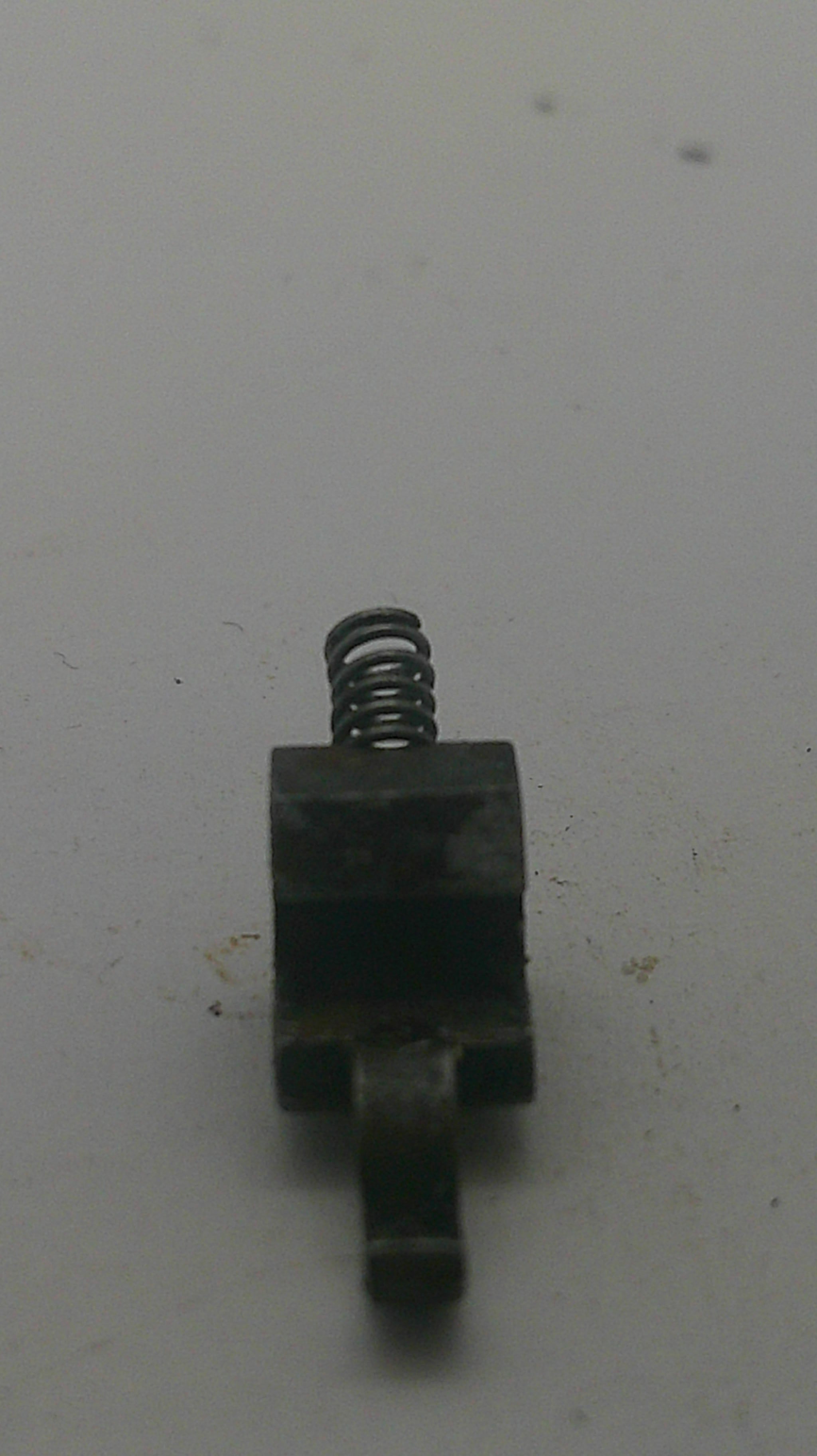 A small dark metal mechanical component with a coiled spring, photographed on a plain off-white surface.