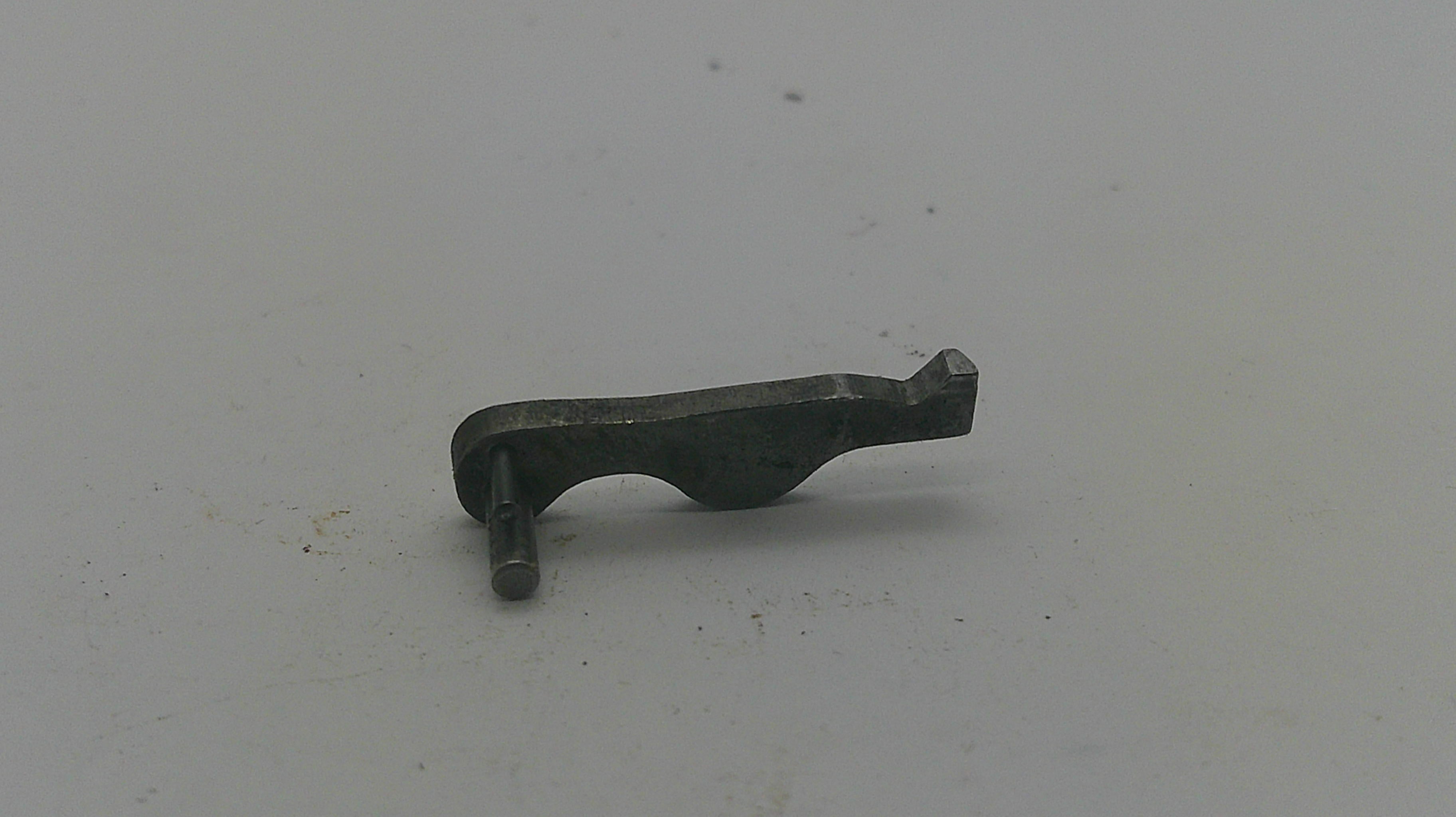 A small dark metal mechanical part or pin with a curved body and flat tip, photographed on a plain light gray surface.