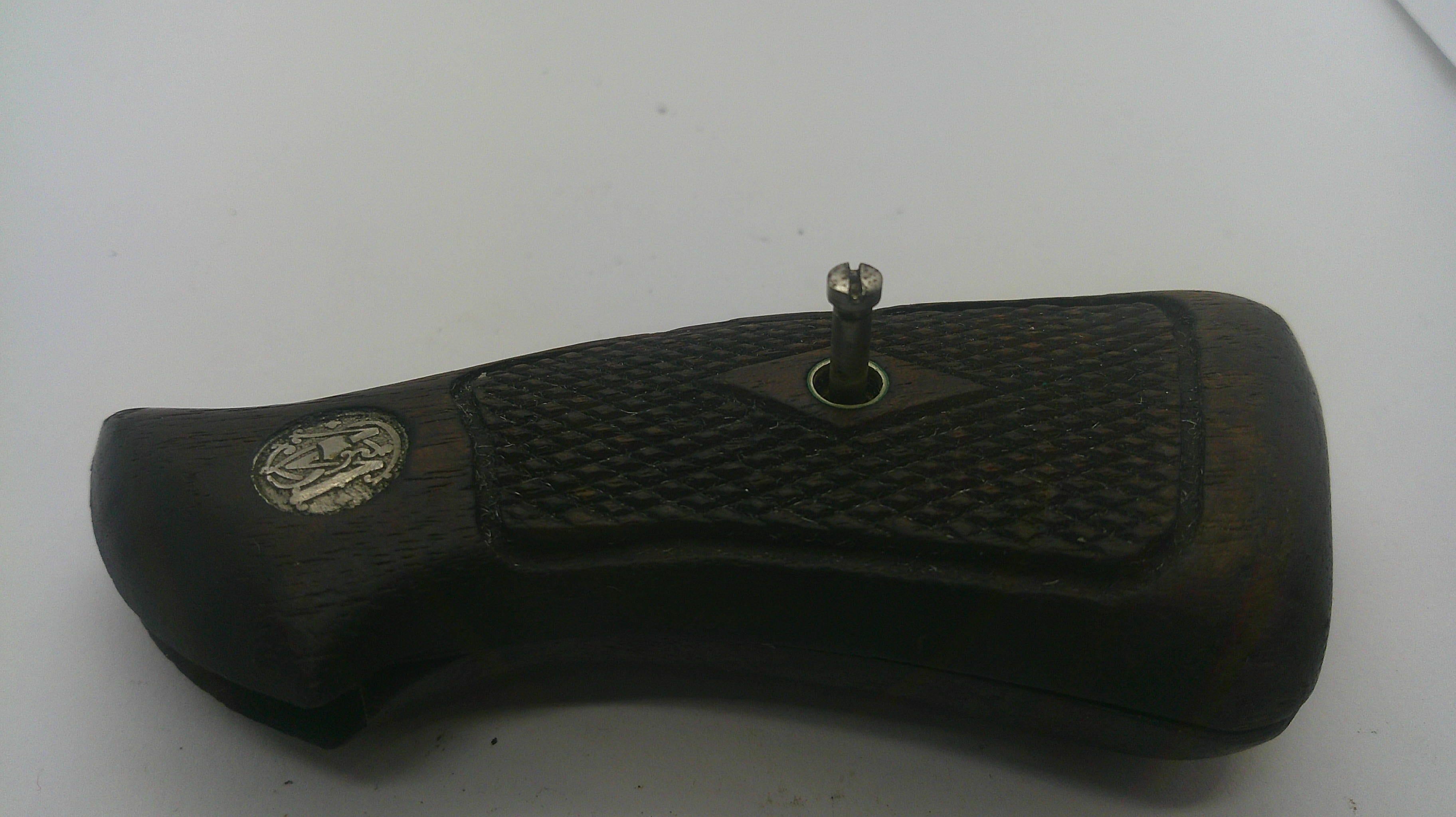A close-up of a dark brown wooden object with a textured grip pattern, a small silver metal knob on top, and a circular silver emblem on the left side, photographed against a plain light background.