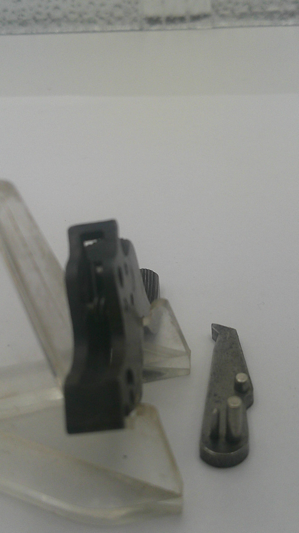 A close-up of a small black plastic mechanical component or clip mounted against a white surface, with a separate small metal piece below it.