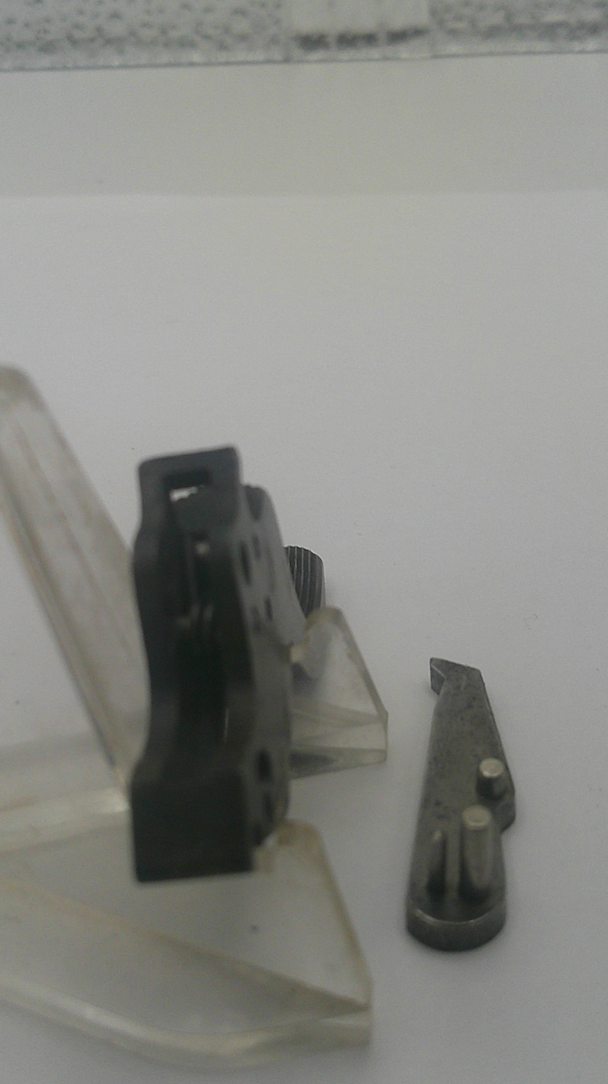 A close-up of a small black plastic mechanical component or clip mounted against a white surface, with a separate small metal piece below it.