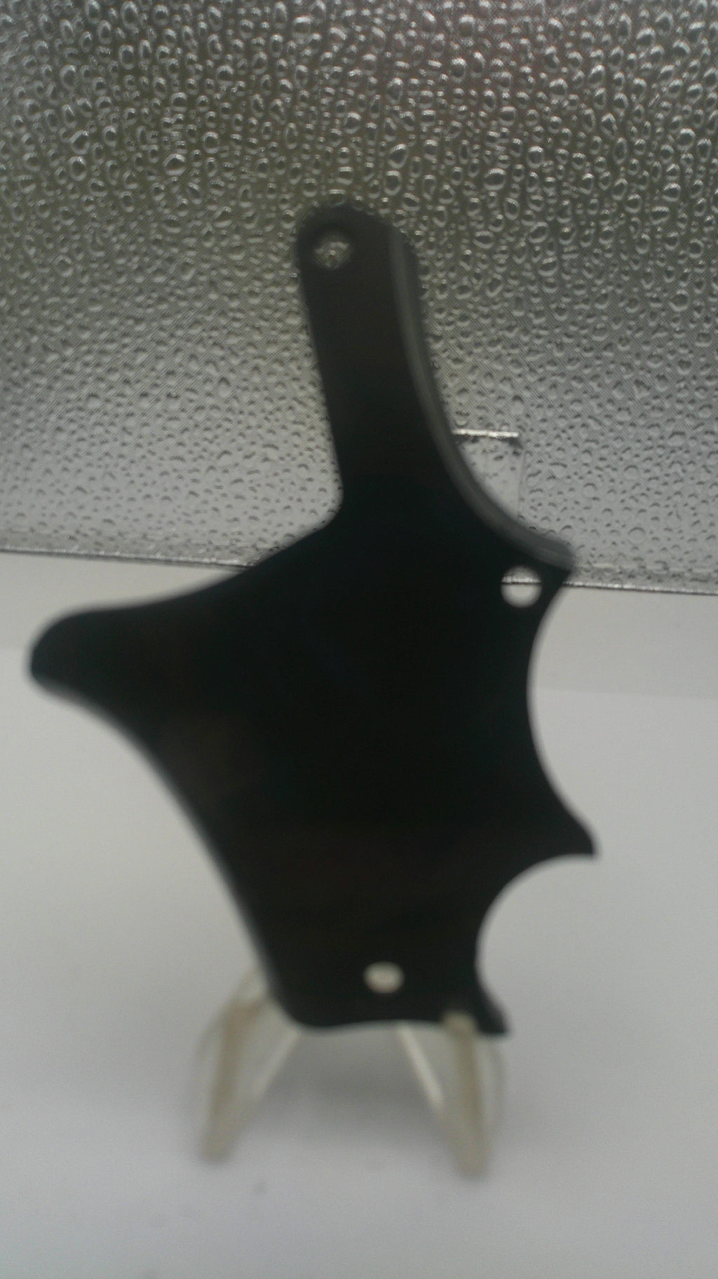 A black plastic mounting bracket or clip with two small silver metal pins, shown against a split background of a white wall and a textured gray surface.