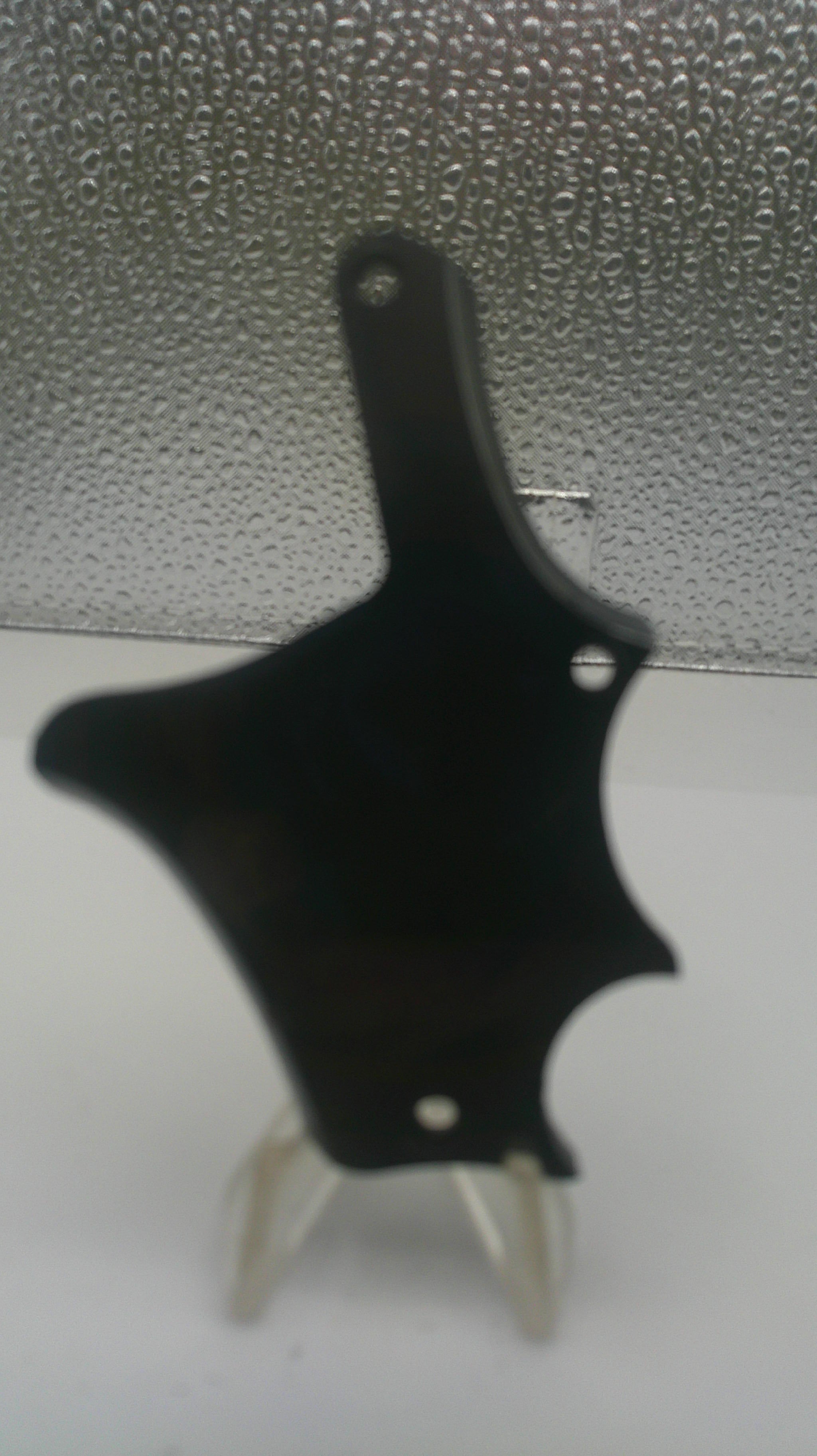 A black plastic mounting bracket or clip with two small silver metal pins, shown against a split background of a white wall and a textured gray surface.