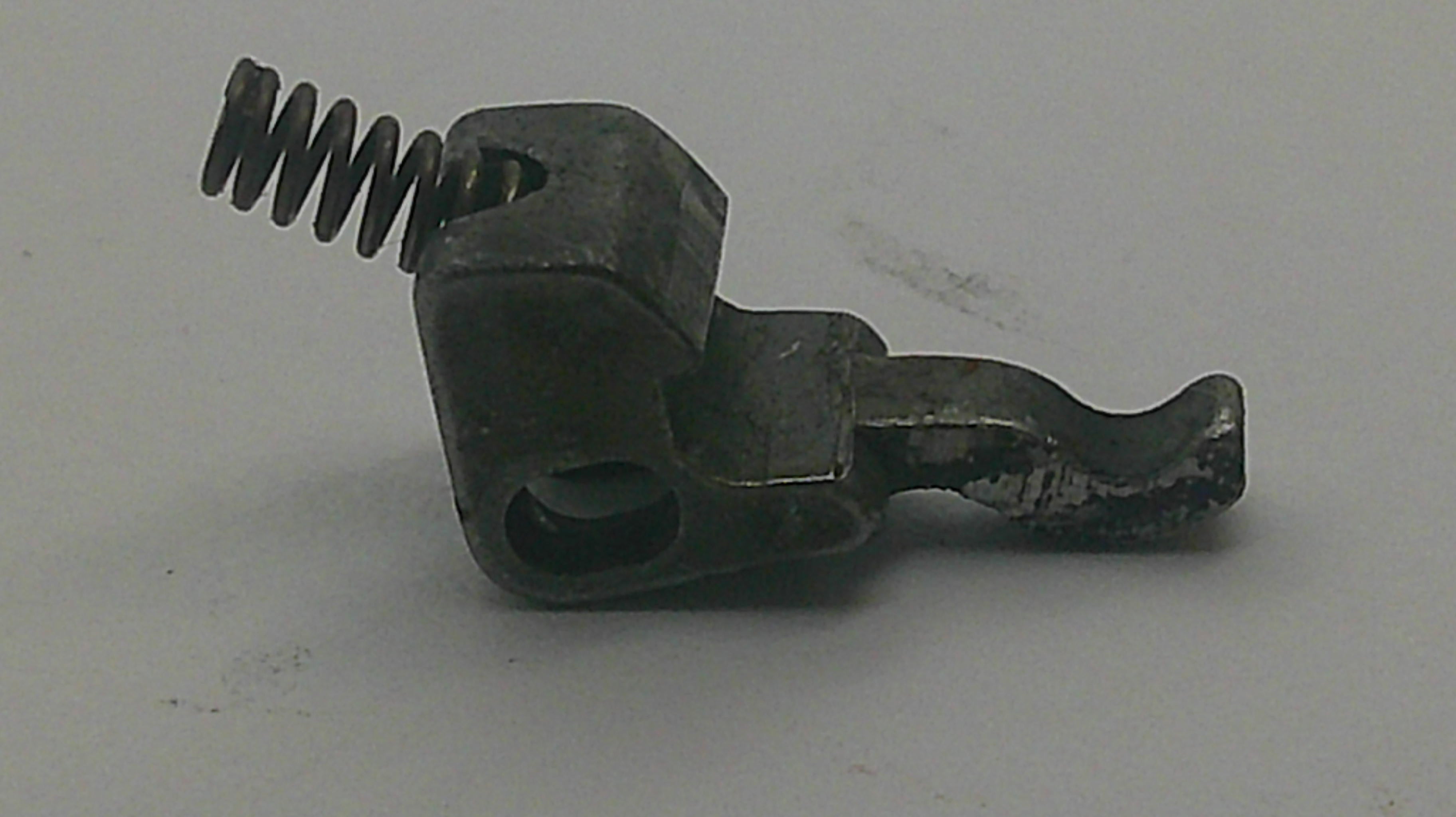 A small dark metal mechanical component with a coiled spring and hook-like arm, photographed on a plain light gray background.