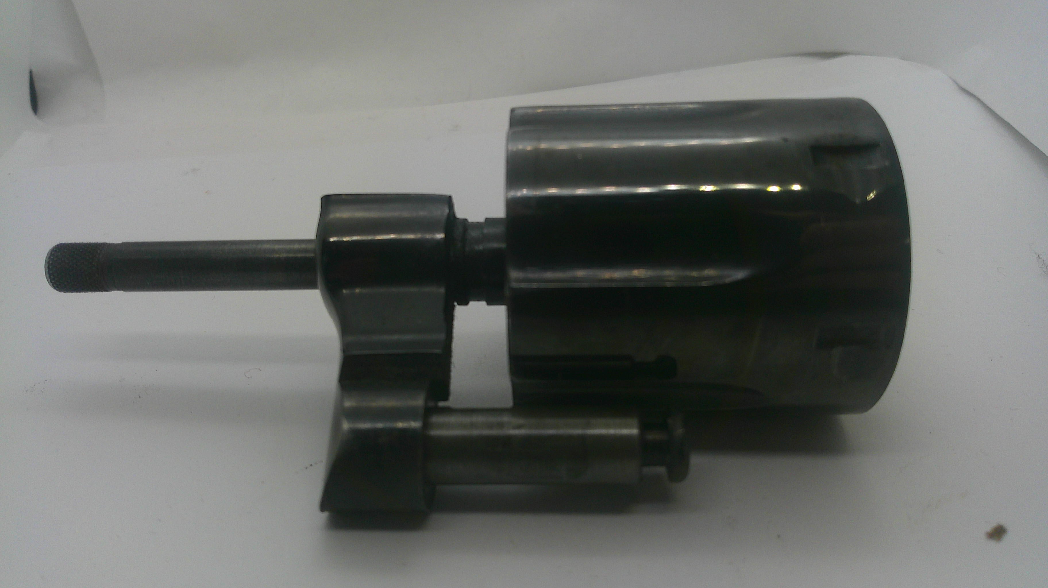 A dark metallic mechanical component with a cylindrical body, a short protruding rod, and a smaller attached rod, photographed on a plain light background.