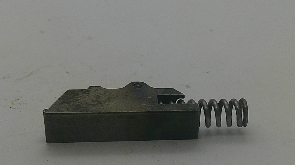 A small dark metal mechanical component with a rectangular block and a coiled spring attached on the right side, photographed on a plain light background.