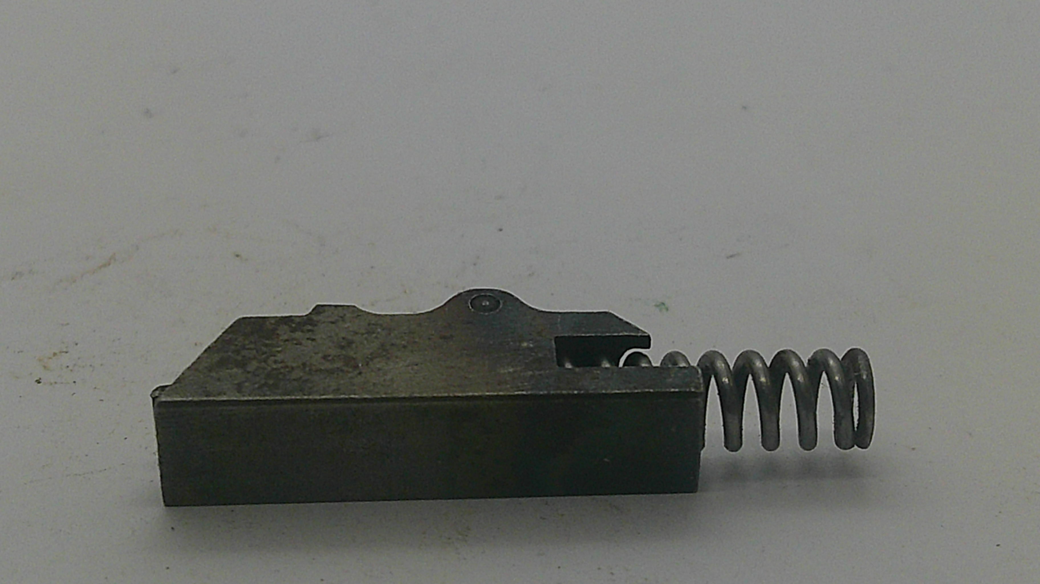 A small dark metal mechanical component with a rectangular block and a coiled spring attached on the right side, photographed on a plain light background.