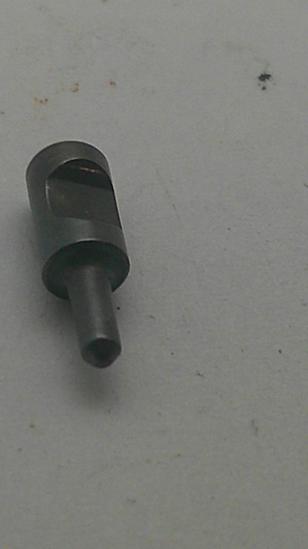A small dark metallic cylindrical tool or pin with a slotted side, photographed on a plain light gray surface.