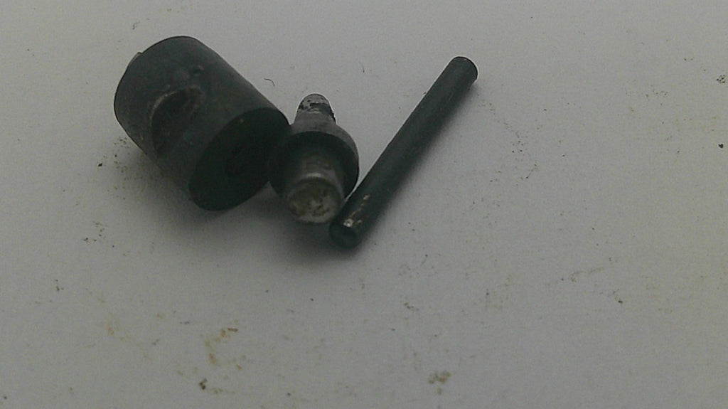 A small set of dark metal mechanical parts, including a cylindrical black component, a silver metal pin, and a dark rod, placed on a light gray surface.