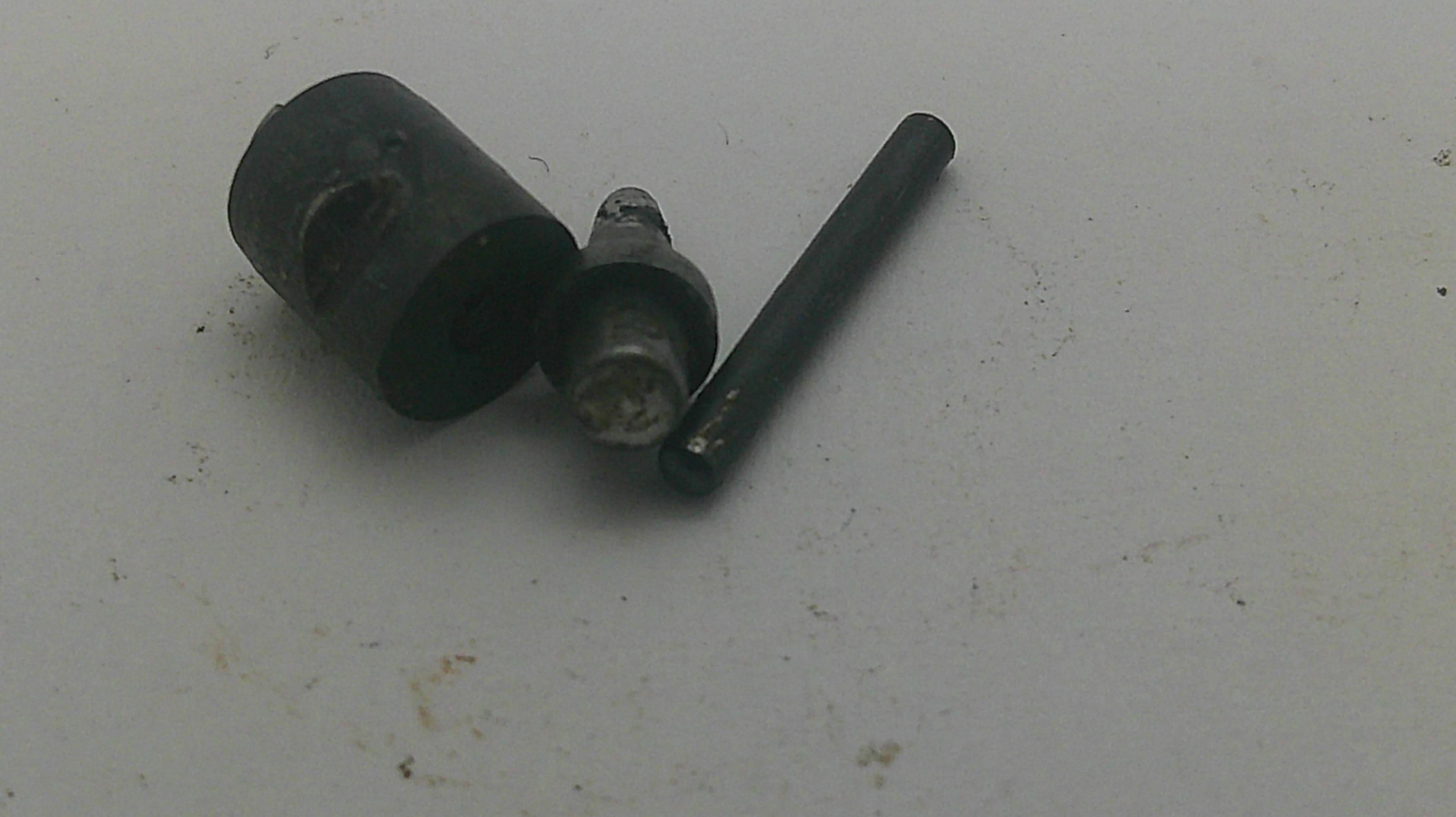 A small set of dark metal mechanical parts, including a cylindrical black component, a silver metal pin, and a dark rod, placed on a light gray surface.