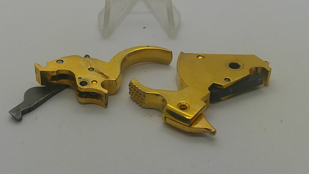 Two gold-finished metal firearm trigger components with a small dark metal piece and a clear plastic display stand on a light gray background.