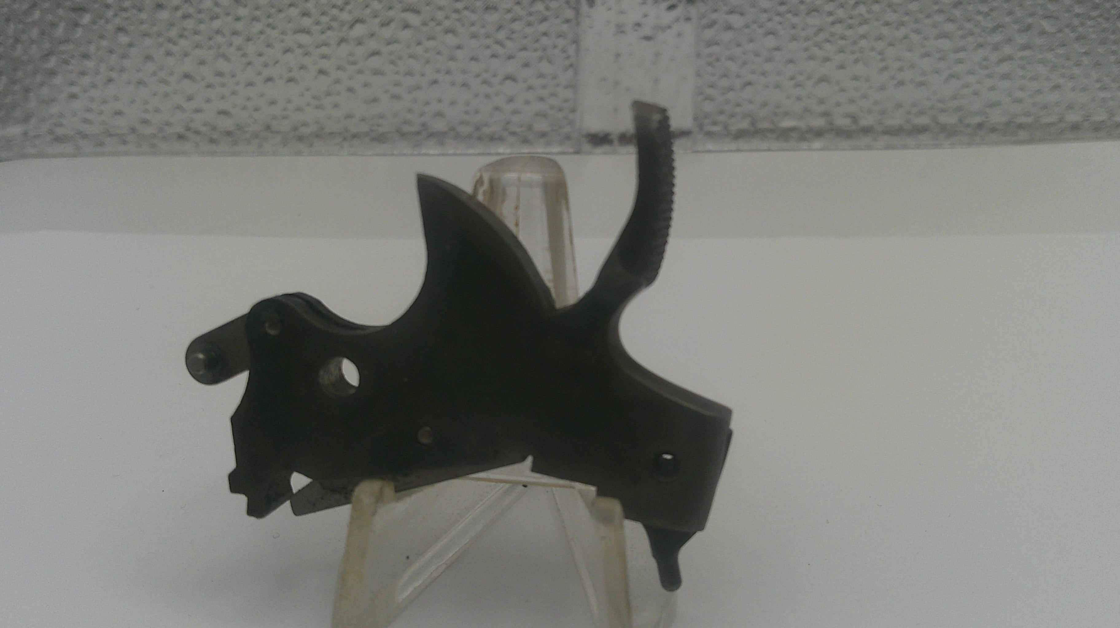 A black metal trigger assembly or lock component displayed on a clear plastic stand against a white surface and textured gray wall background.