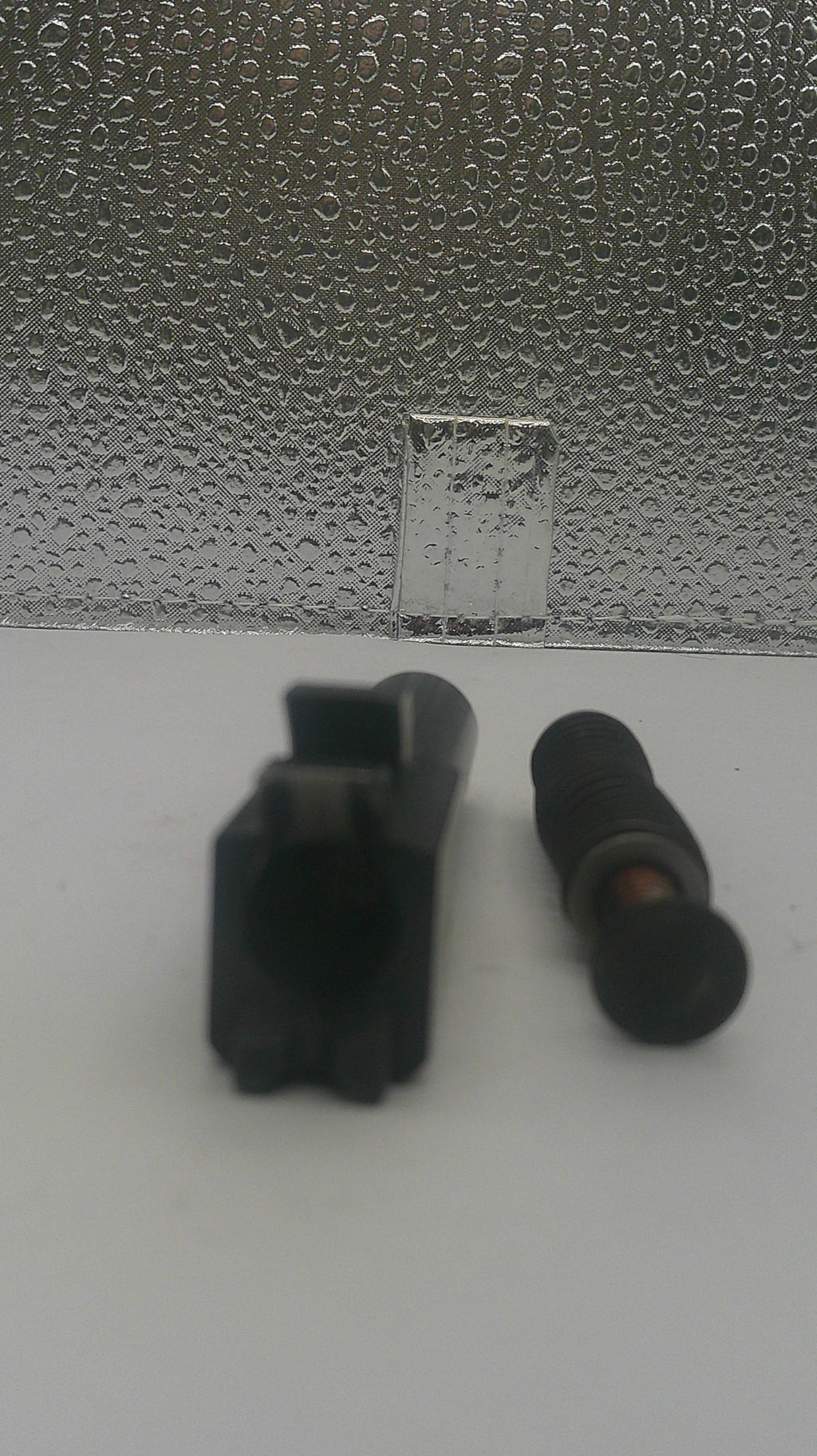 Two small black plastic automotive or appliance parts, one rectangular with a circular opening and one cylindrical with a threaded tip, shown on a white surface next to a textured silver panel.
