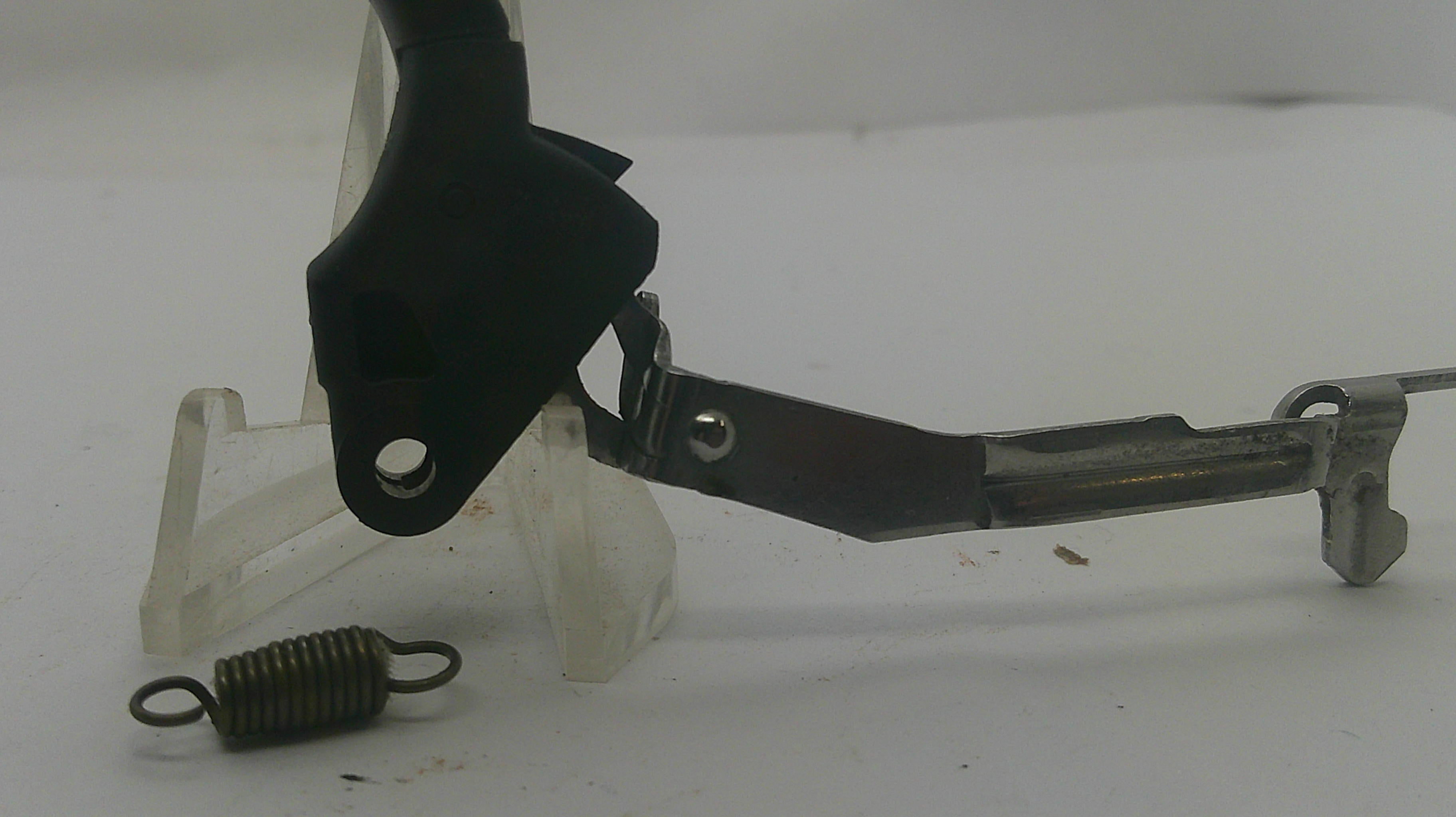 A metal mechanical lever assembly with a black plastic mounting piece, a small coiled spring, and a clear plastic display stand on a white background.
