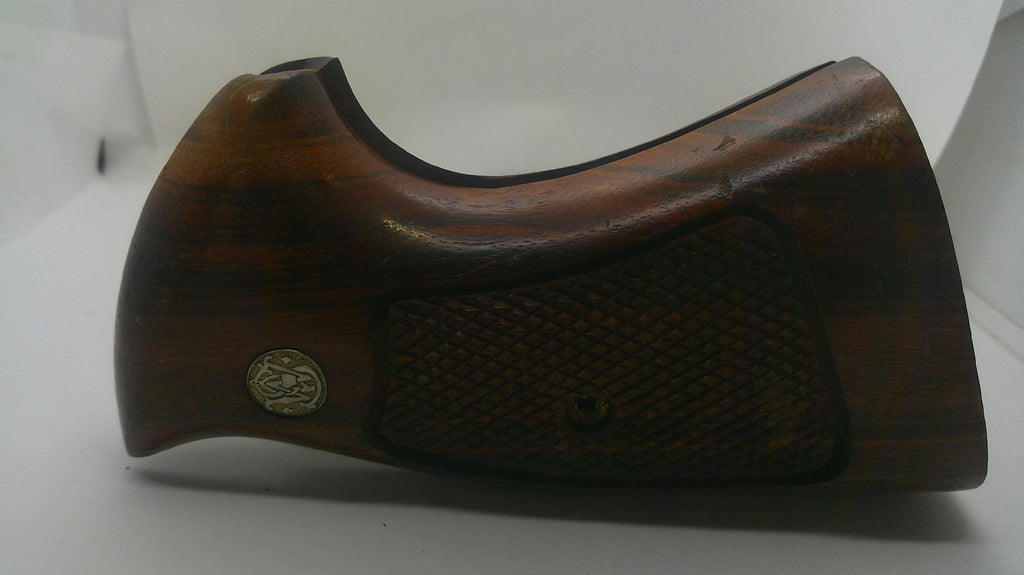 A dark brown wooden gunstock grip or handle component with a textured checkered pattern and a small circular metal emblem, shown on a plain white background.