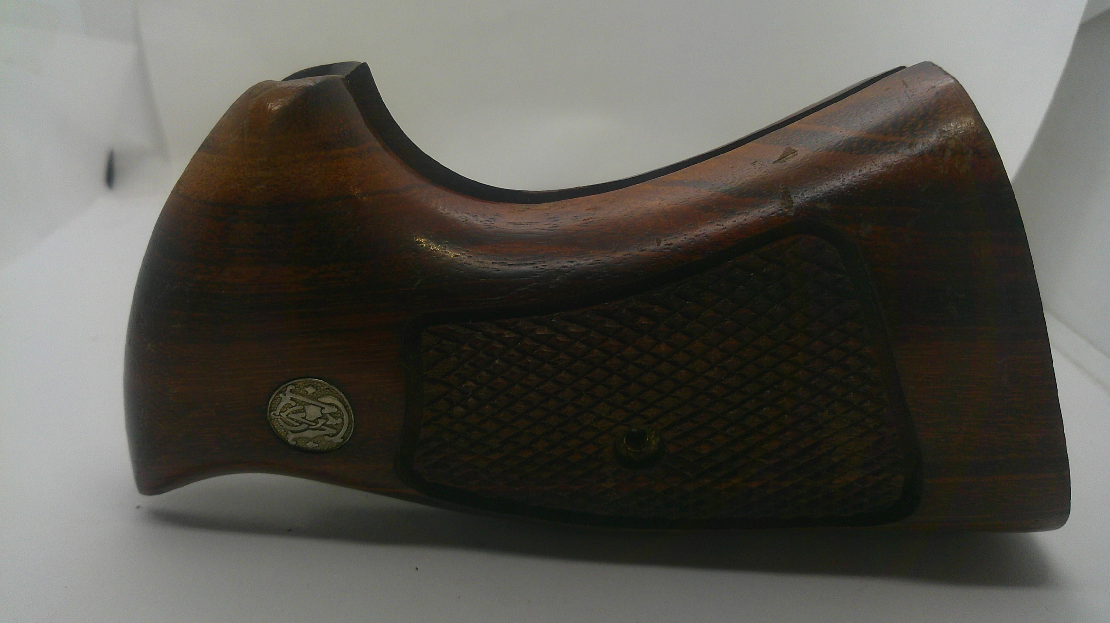 A dark brown wooden gunstock grip or handle component with a textured checkered pattern and a small circular metal emblem, shown on a plain white background.
