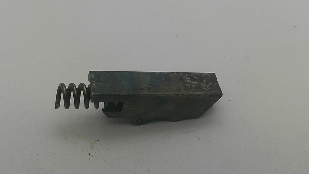 A small dark metal mechanical component with a coiled spring attached to one end, photographed on a plain off-white background.