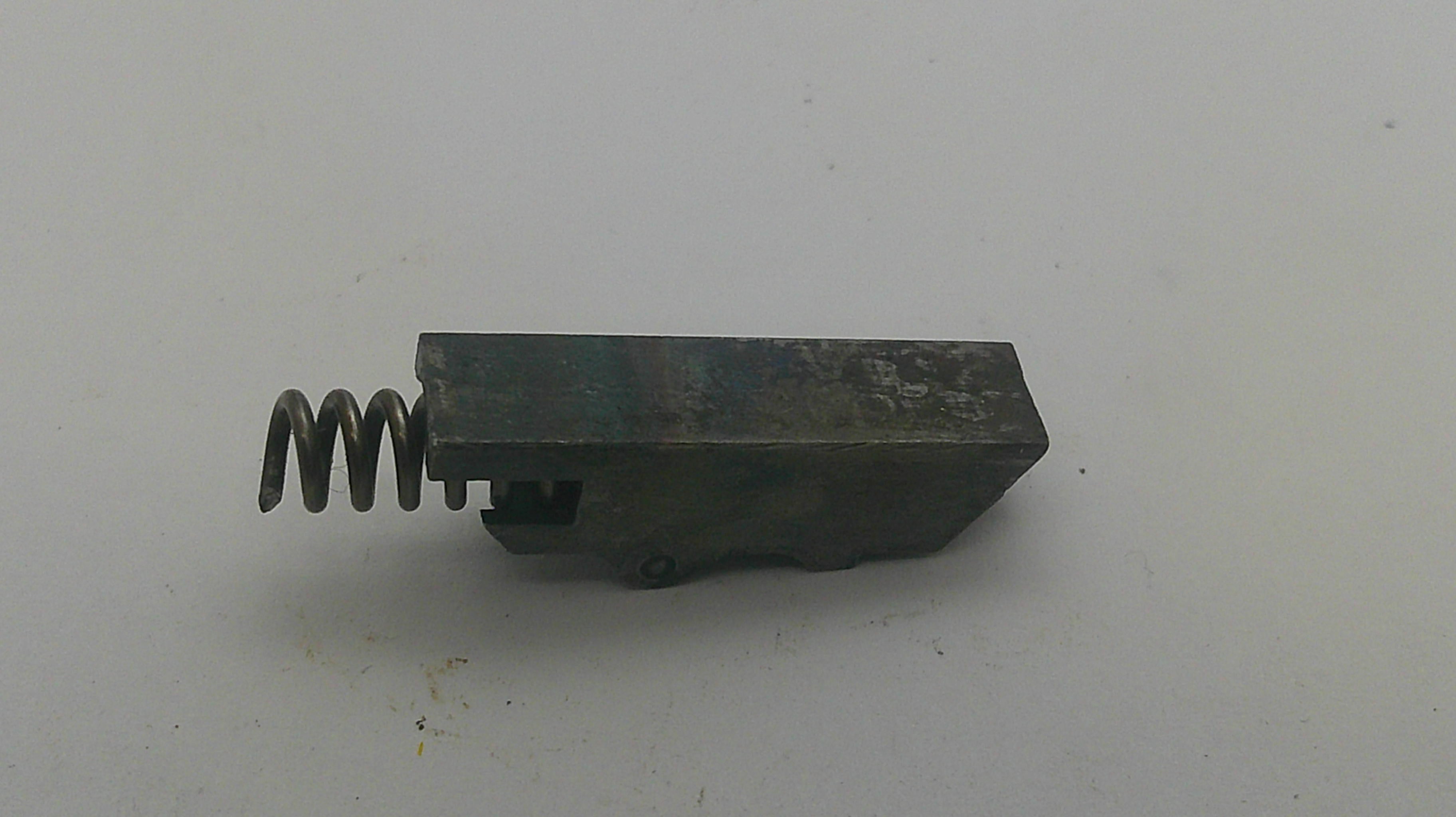 A small dark metal mechanical component with a coiled spring attached to one end, photographed on a plain off-white background.