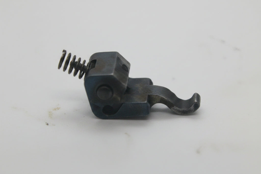 A small dark metal mechanical component with a spring, shown on a plain off-white background.