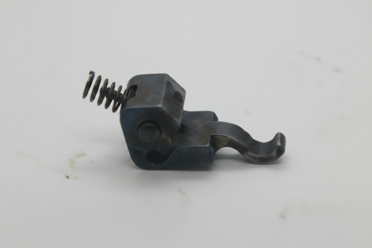 A small dark metal mechanical component with a spring, shown on a plain off-white background.