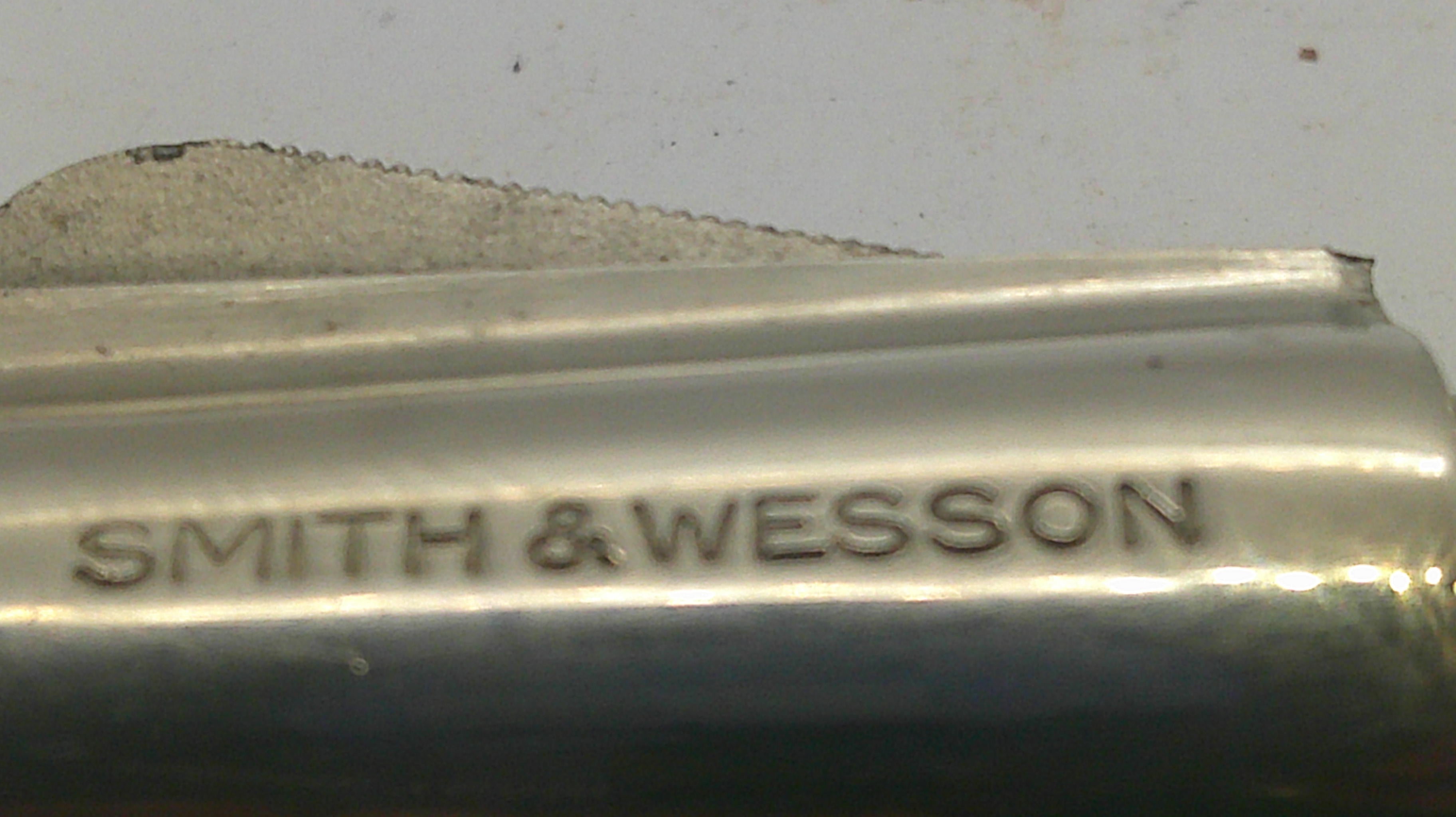 Close-up of a metallic cylindrical object with the brand name 'SMITH & WESSON' engraved on it, against a light gray background.