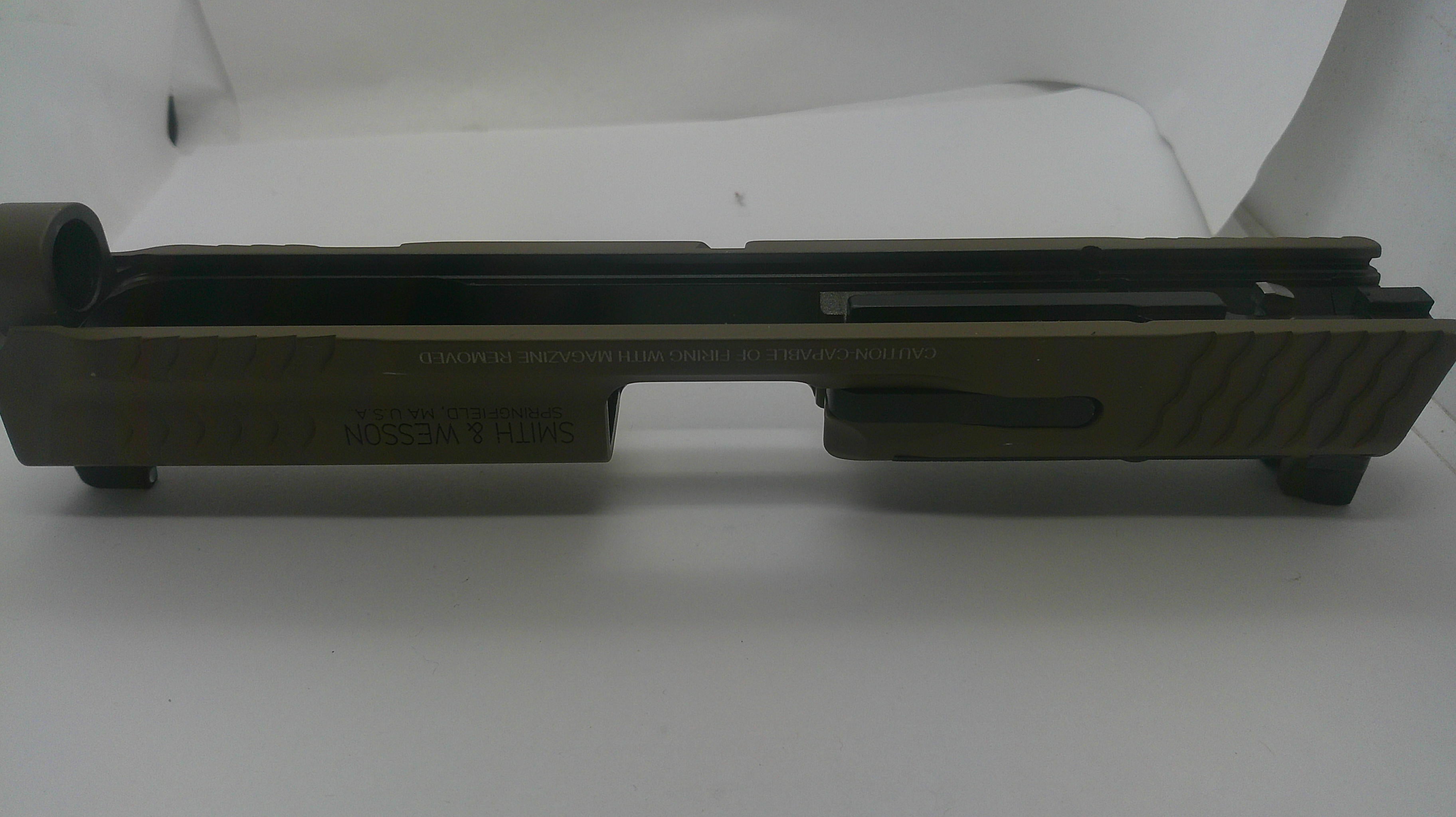 An olive drab green metal receiver or slide component with black rubber boots and a black textured grip, photographed against a plain white background.