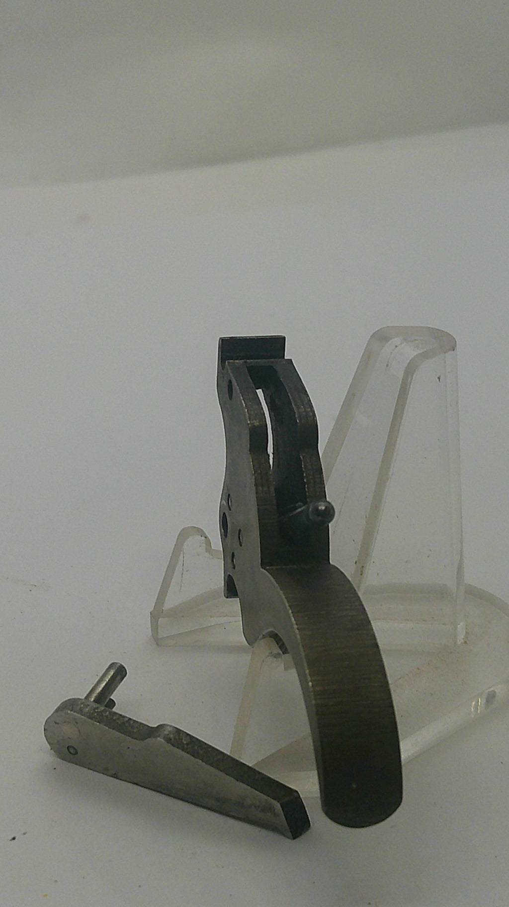 A close-up of a dark metal mechanical component or tool mounted on a clear plastic display stand against a plain light background.
