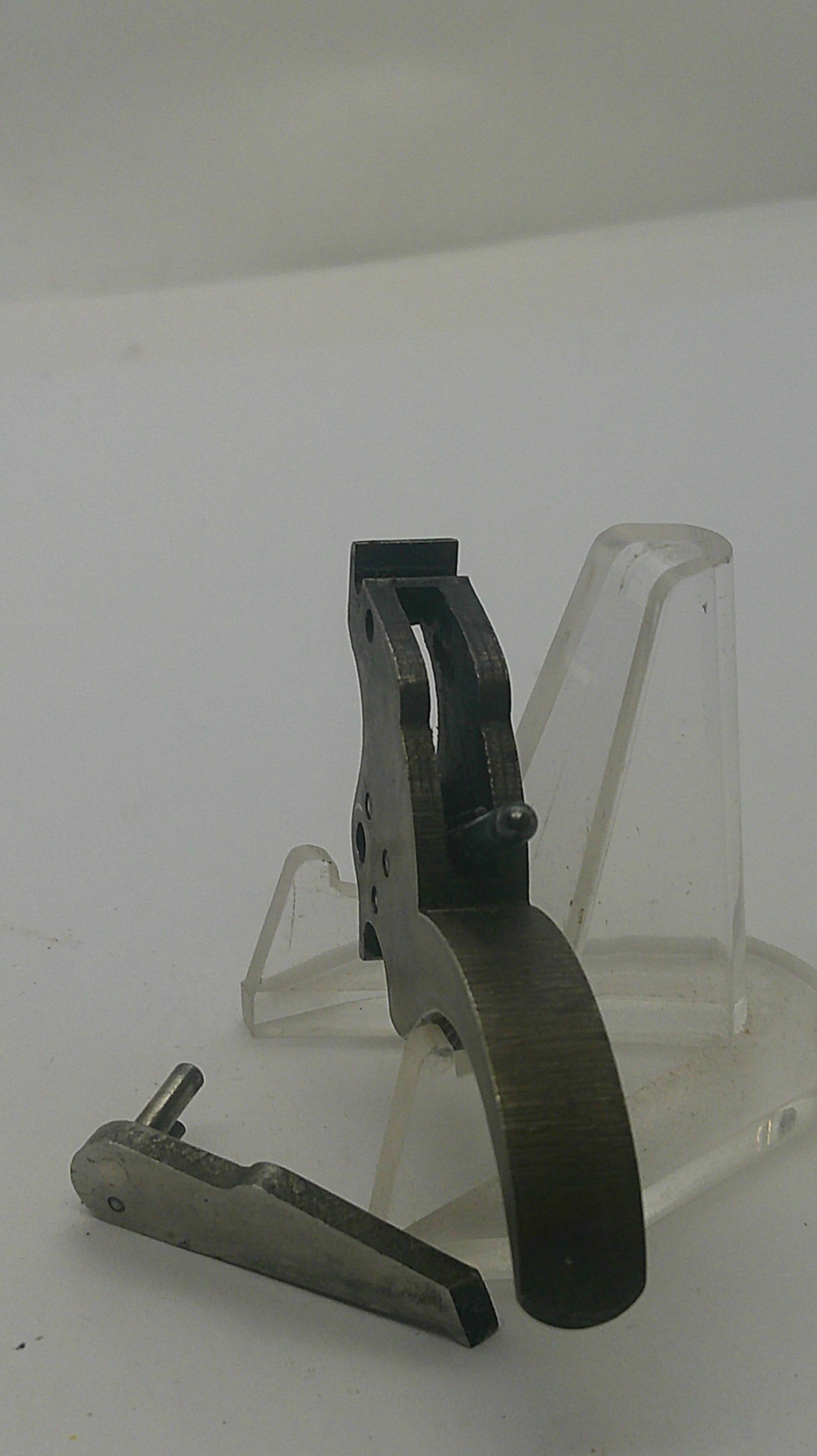 A close-up of a dark metal mechanical component or tool mounted on a clear plastic display stand against a plain light background.