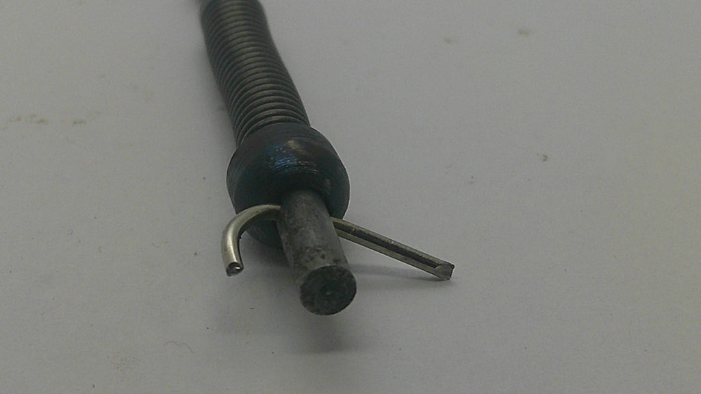 A close-up of a flexible metal tube with a coiled spring-like section, a dark round connector, and two small metal prongs or clips on a plain light background.