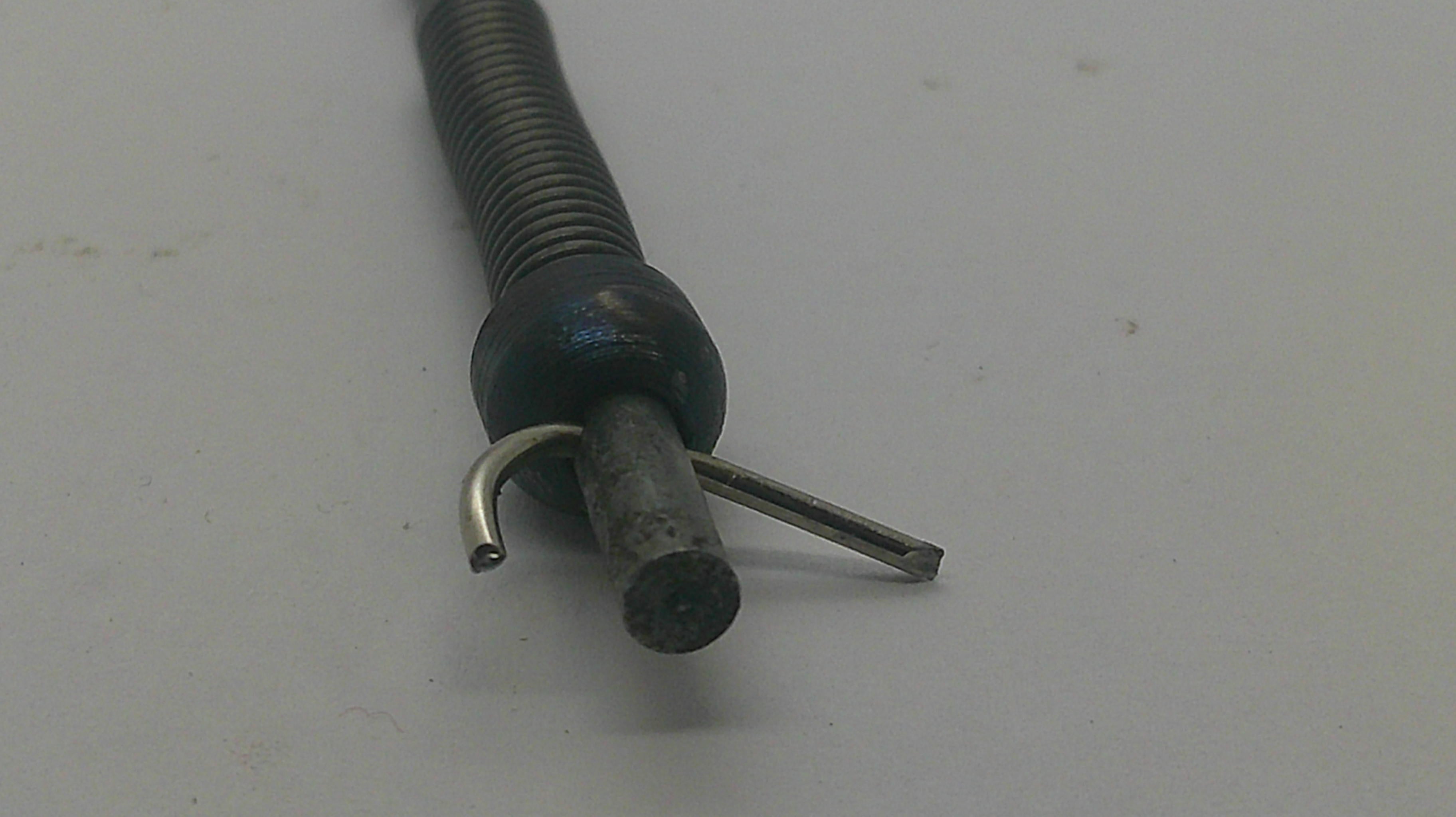 A close-up of a flexible metal tube with a coiled spring-like section, a dark round connector, and two small metal prongs or clips on a plain light background.