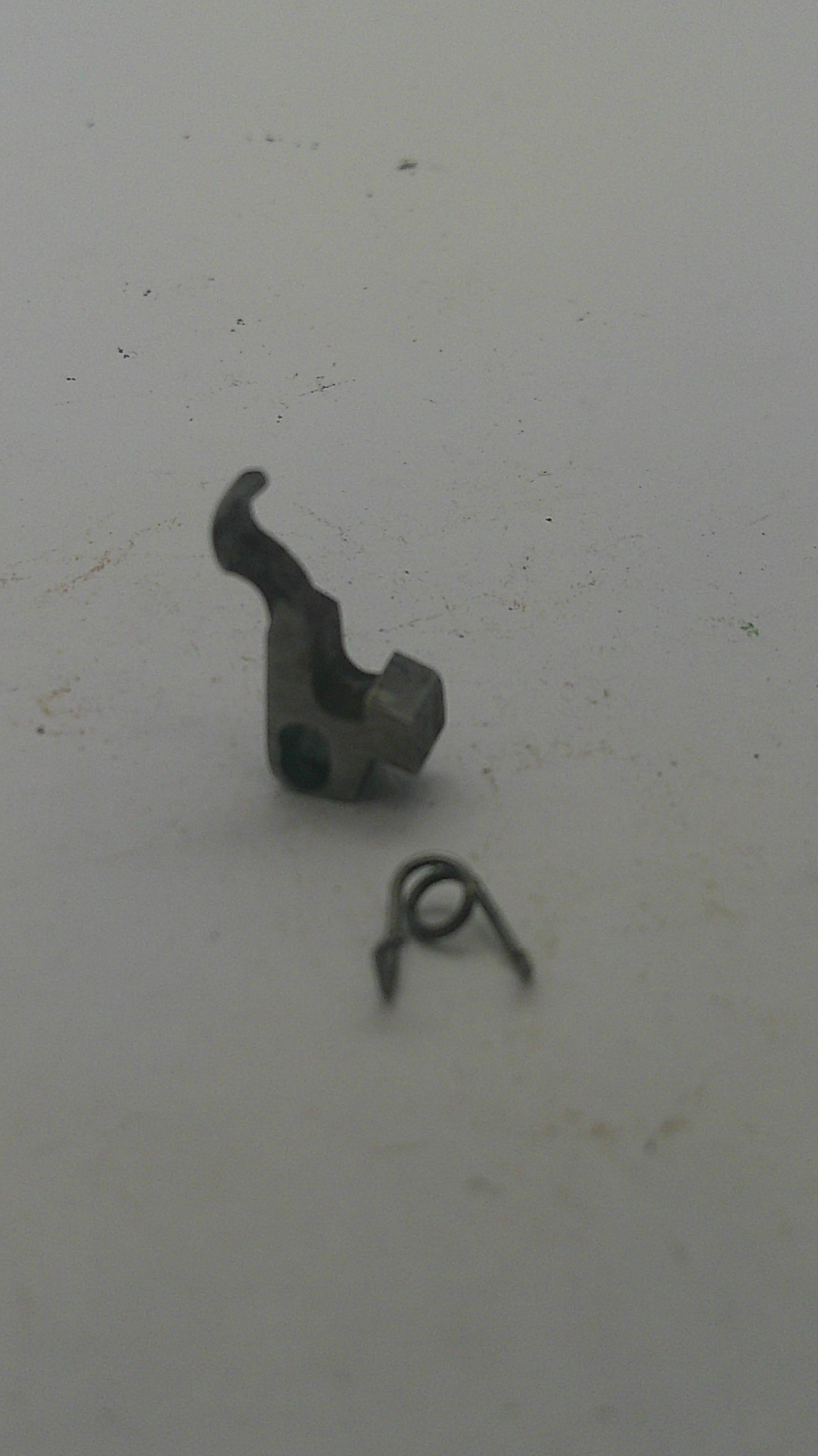 A small dark metal mechanical component with a curved hook-like shape, shown next to a separate small spring on a plain off-white surface.