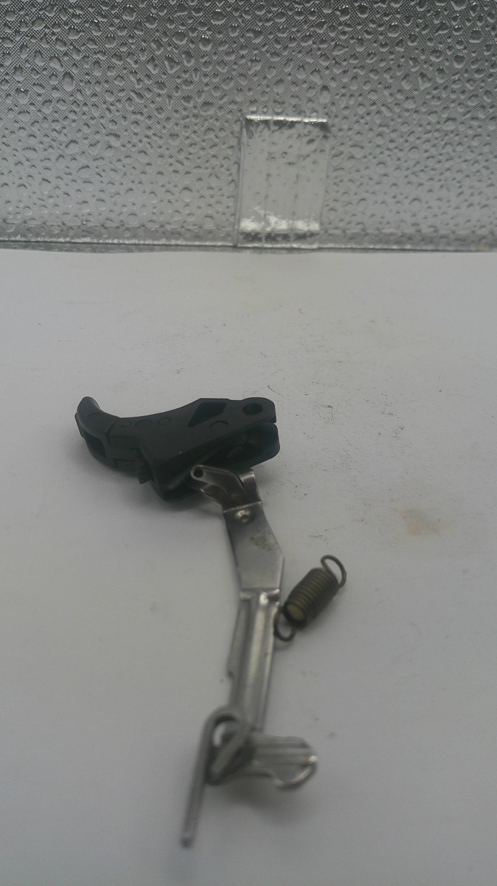 A small mechanical part consisting of a silver metal lever and a black plastic component, placed on a light gray surface next to a textured gray panel.