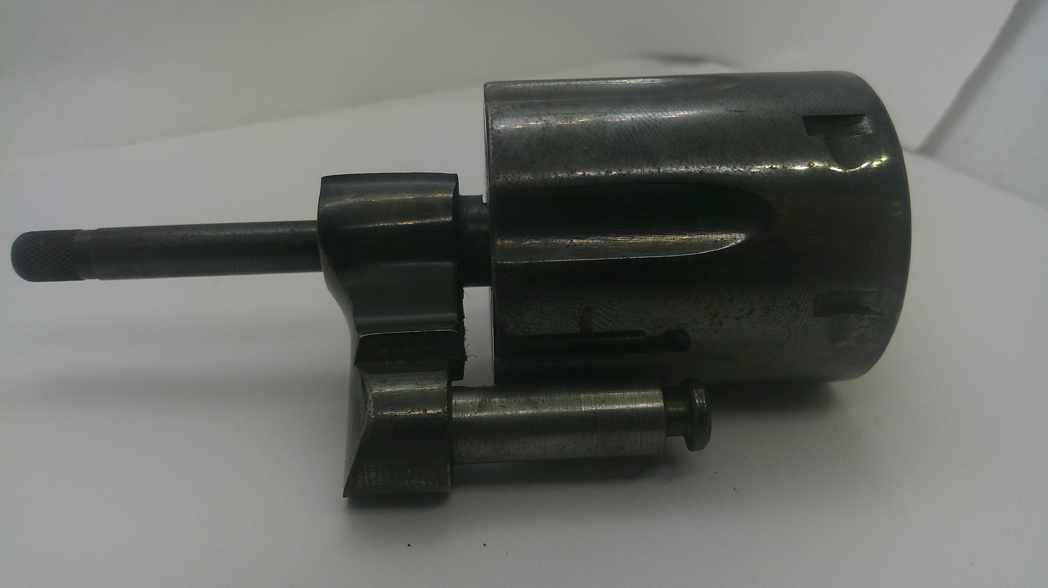 A dark metallic mechanical component with a cylindrical body, a protruding rod, and a small attached cylindrical piece, photographed on a plain light background.