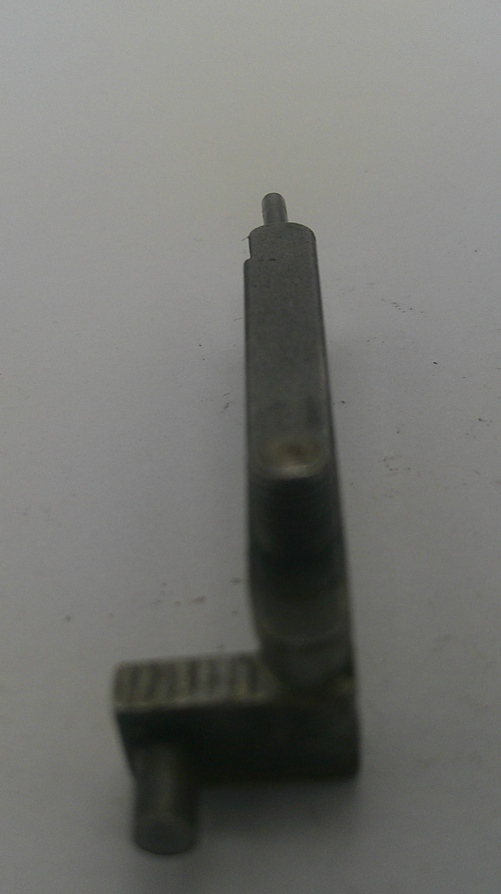 A small dark metal mechanical part or tool component with a flat rectangular body and a pointed tip, photographed on a plain light background.