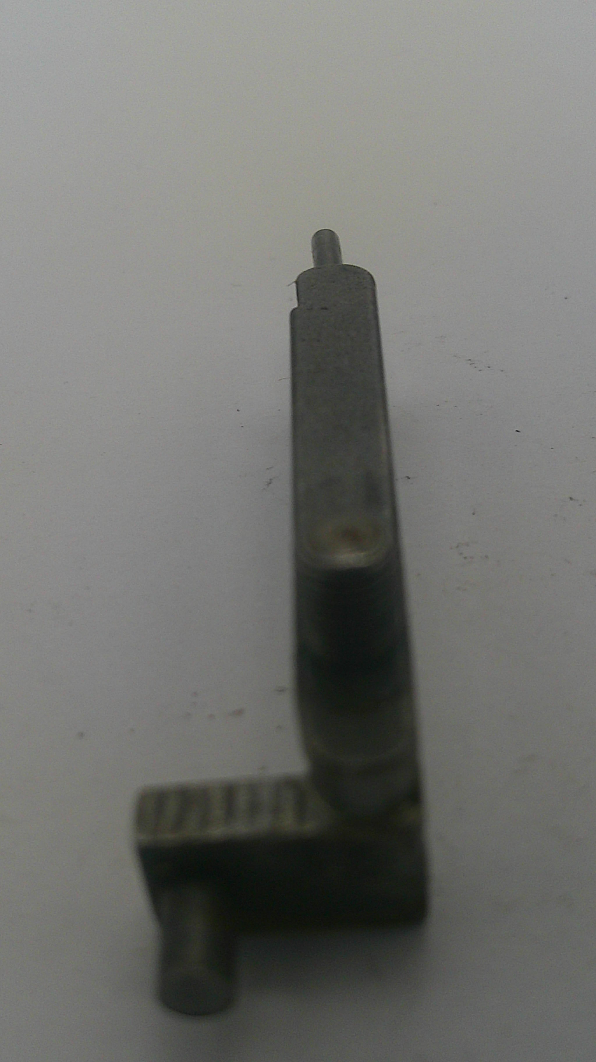 A small dark metal mechanical part or tool component with a flat rectangular body and a pointed tip, photographed on a plain light background.