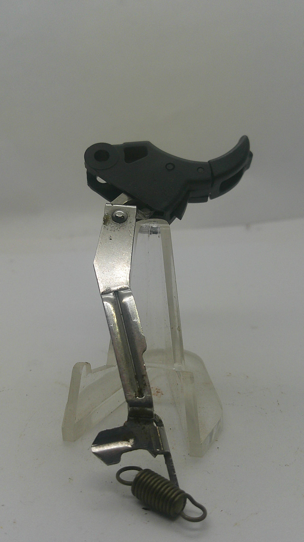 A replacement trigger assembly for a firearm, featuring a black plastic trigger, a silver metal lever, and a black coil spring, mounted on a clear plastic display stand against a plain white background.