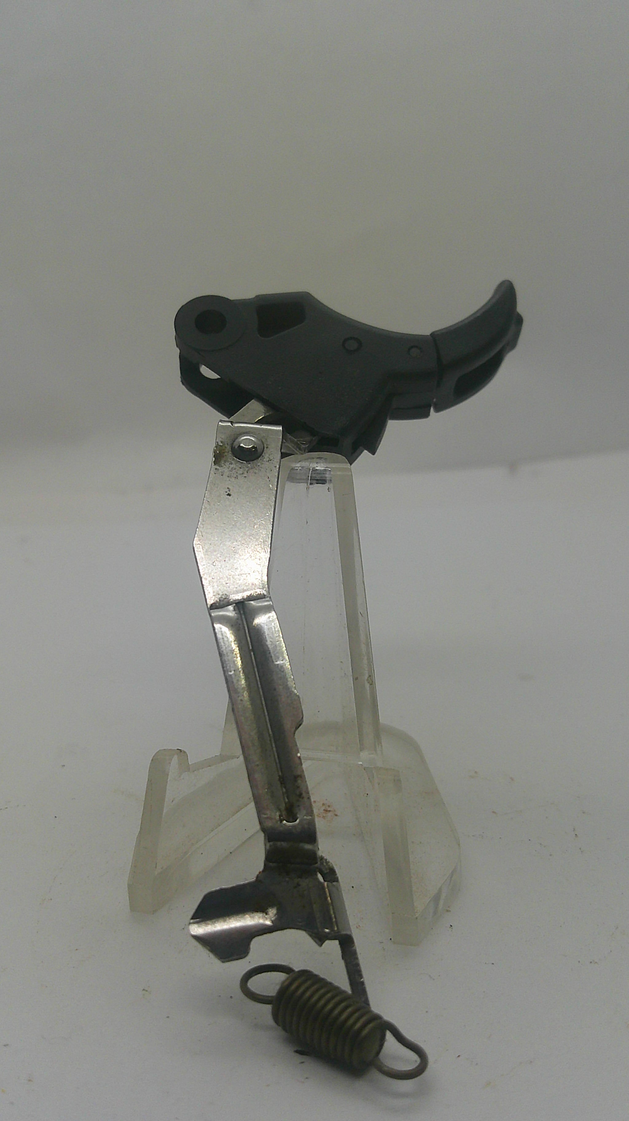 A replacement trigger assembly for a firearm, featuring a black plastic trigger, a silver metal lever, and a black coil spring, mounted on a clear plastic display stand against a plain white background.