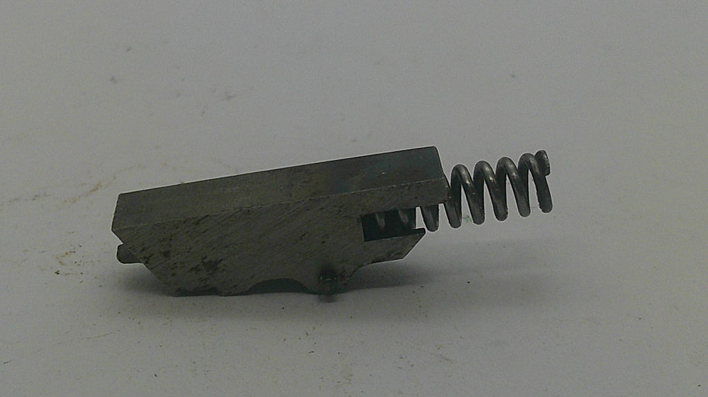 A small metal mechanical component with a rectangular block and a coiled spring, photographed on a plain light background.