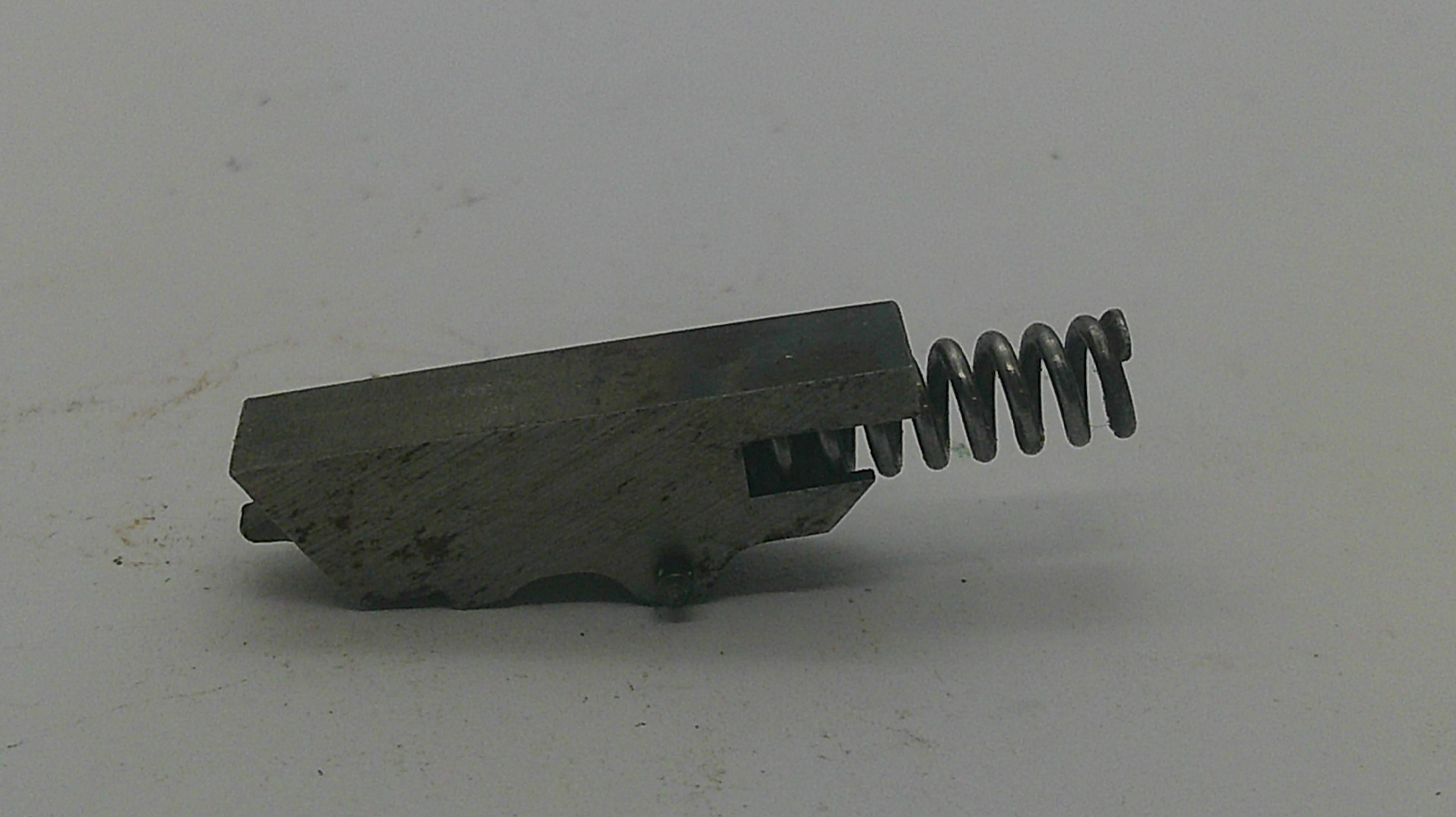 A small metal mechanical component with a rectangular block and a coiled spring, photographed on a plain light background.