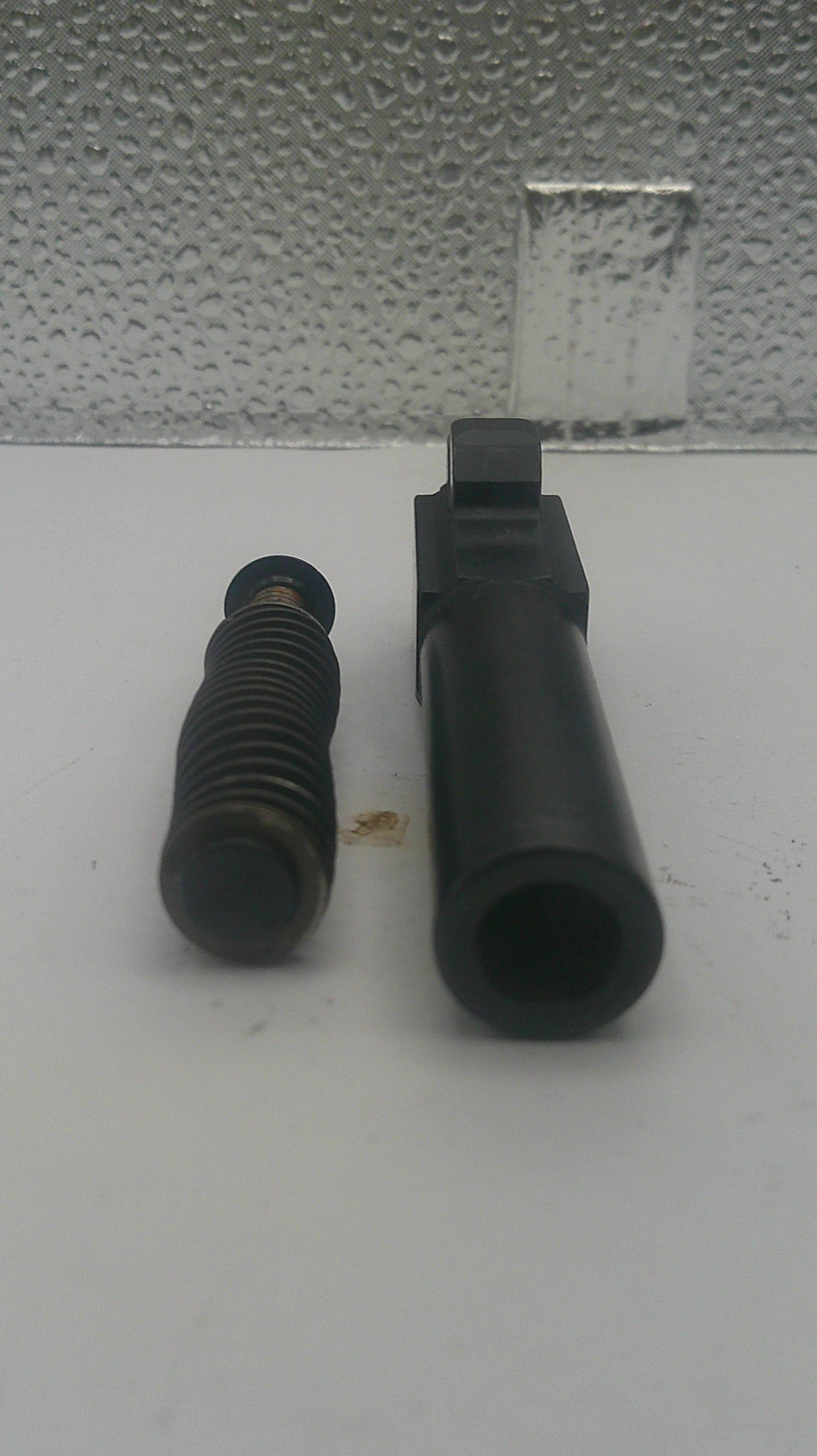 A black metal cylindrical component with a threaded section and a flat mounting tab, photographed on a white surface next to a textured gray wall.