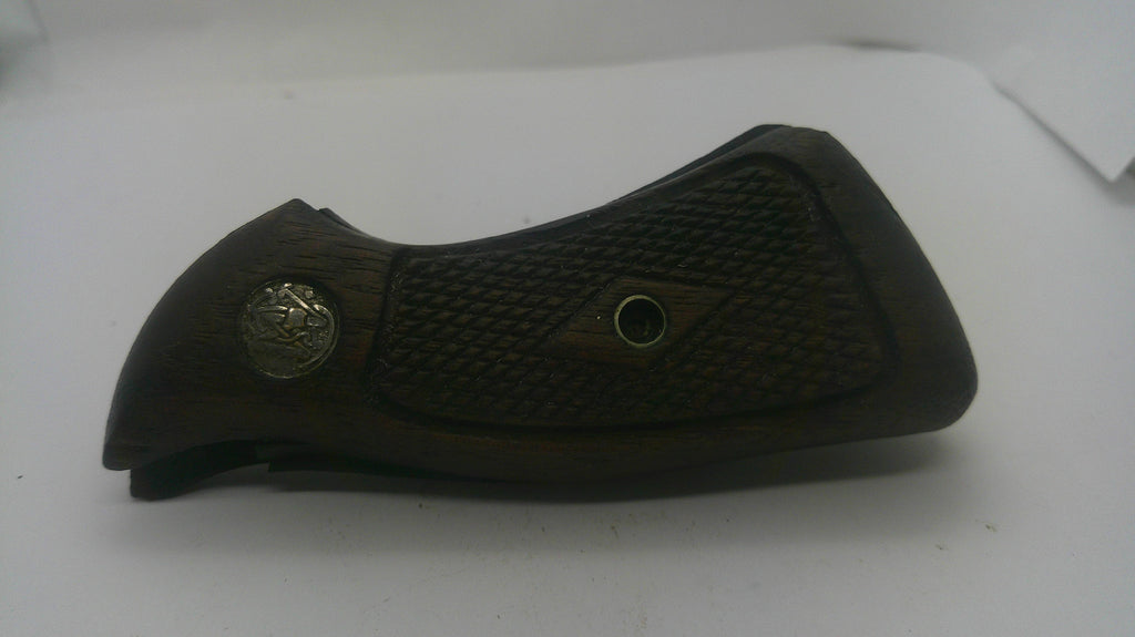 A dark brown wooden gun grip with a textured checkered pattern, featuring a silver-toned metal emblem on the left and a central diamond-shaped grip defect patch.