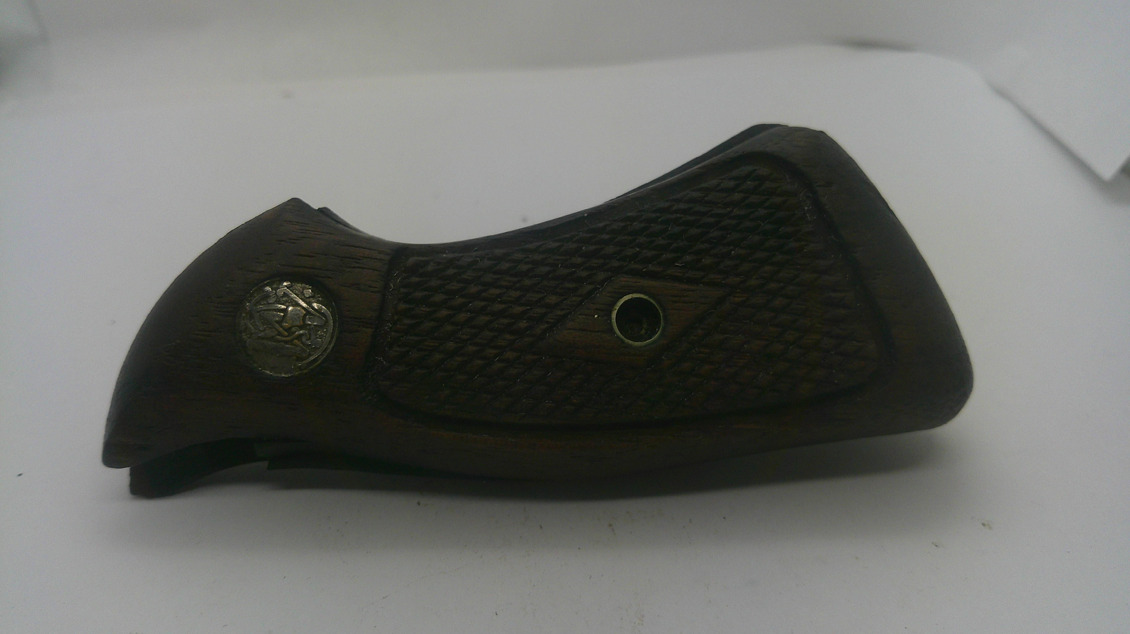 A dark brown wooden gun grip with a textured checkered pattern, featuring a silver-toned metal emblem on the left and a central diamond-shaped grip defect patch.