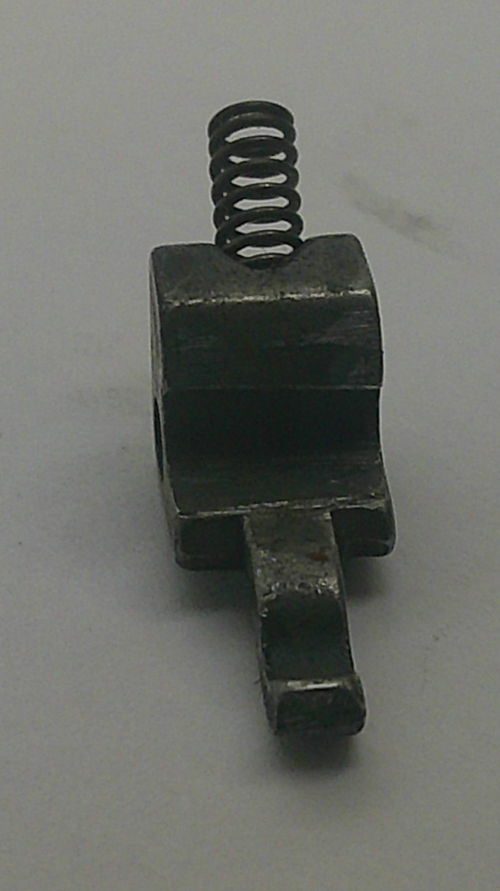 A small dark metal mechanical component with a hexagonal body, a flat tab on one side, and a coiled spring attached to the other side, photographed on a plain light background.