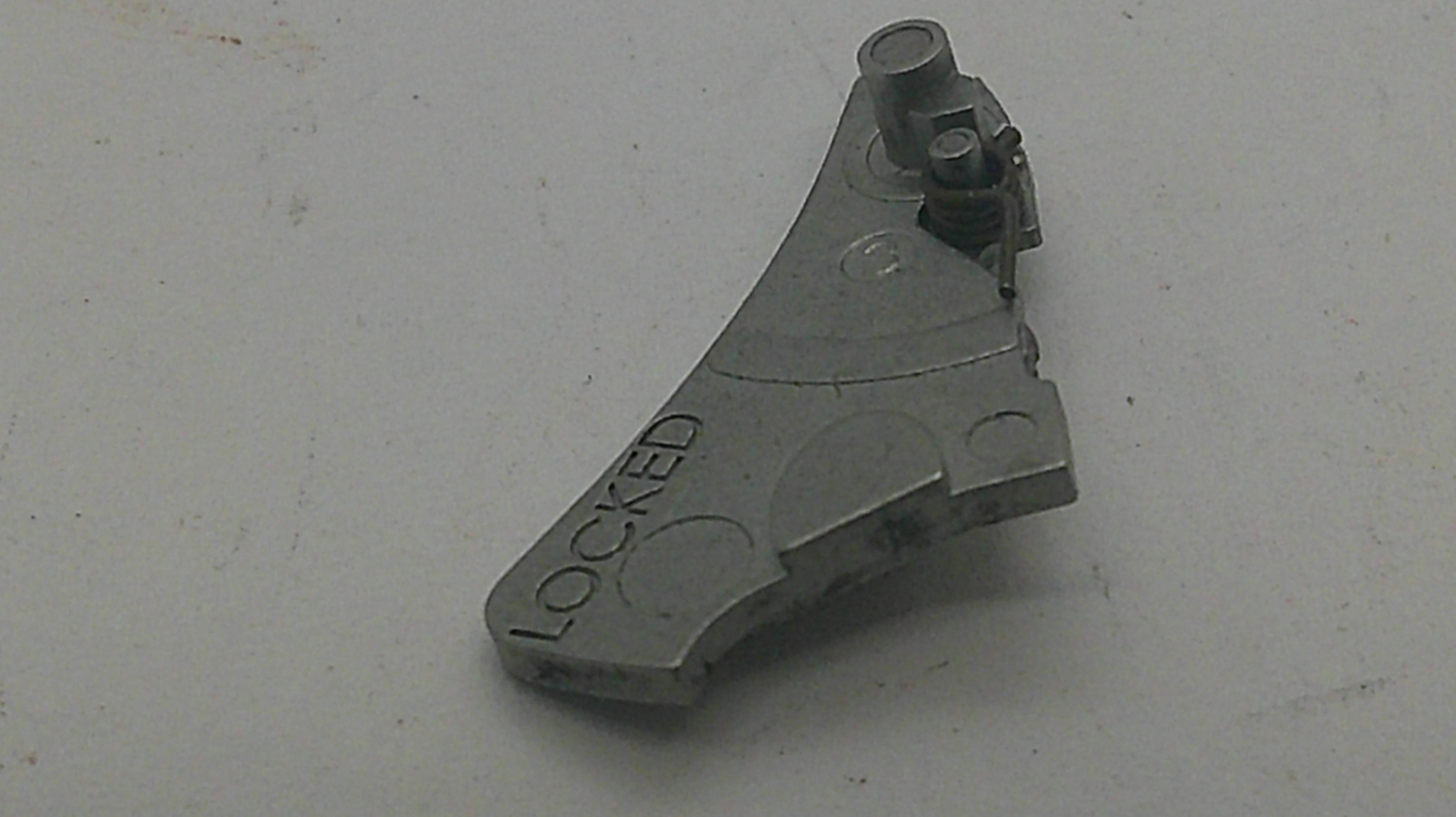 A small gray metal mechanical part with the word 'LOCKED' molded into it, photographed on a plain light surface.