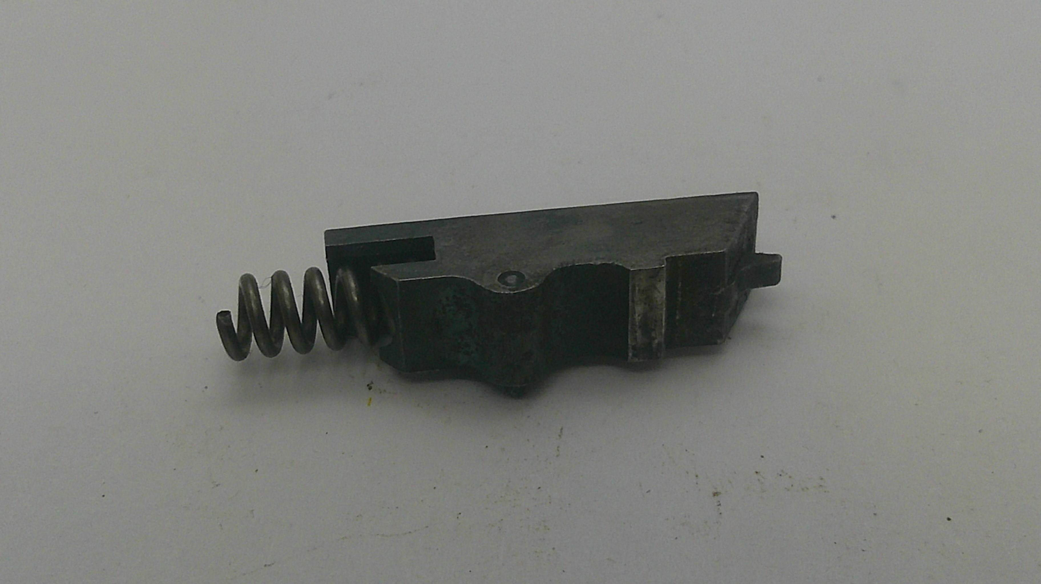 A small dark metal mechanical component with a coiled spring attached, photographed on a plain off-white surface.