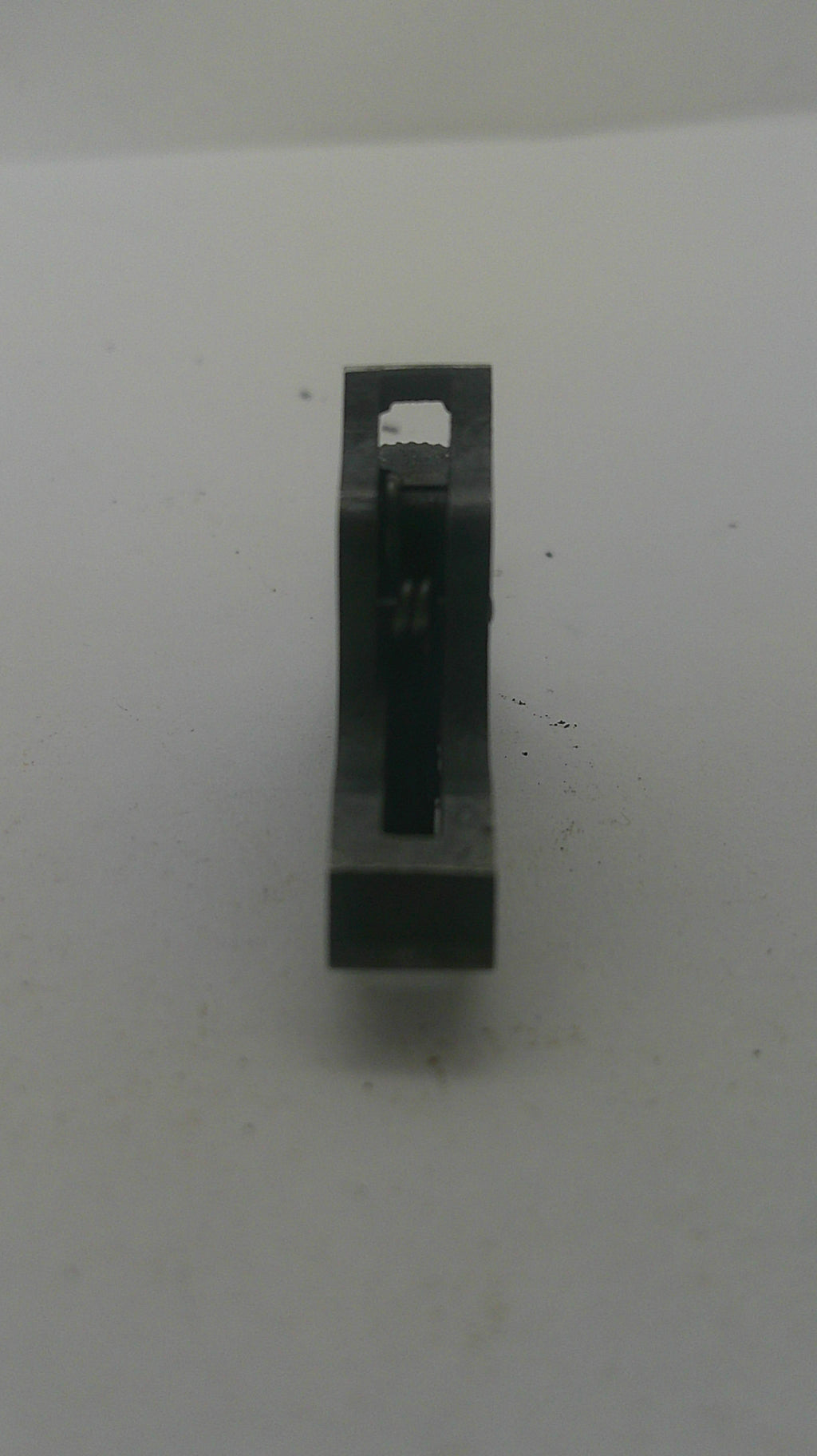 A small black metal rectangular component with a central slot and a small white tab, photographed on a plain off-white surface.