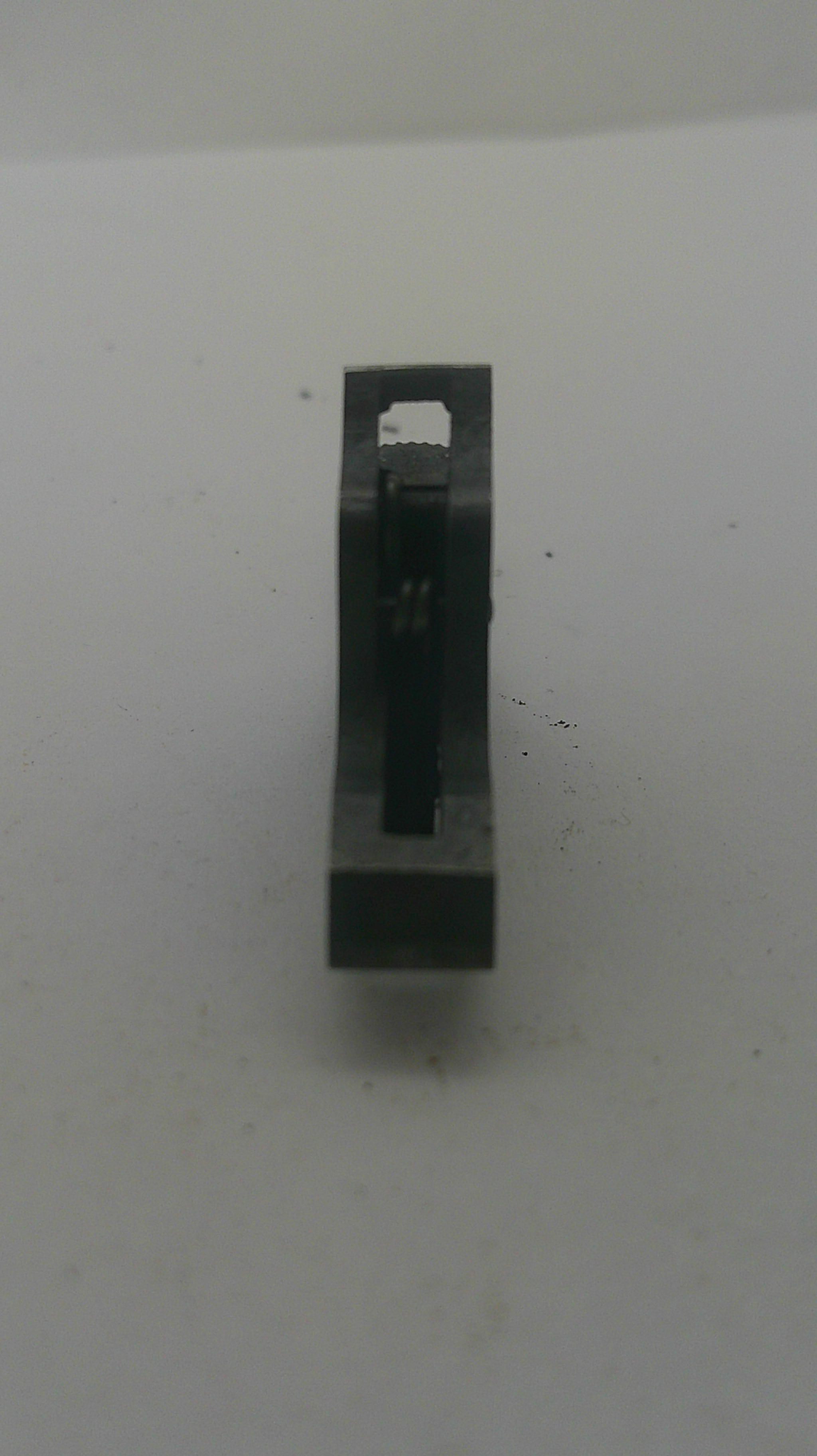 A small black metal rectangular component with a central slot and a small white tab, photographed on a plain off-white surface.