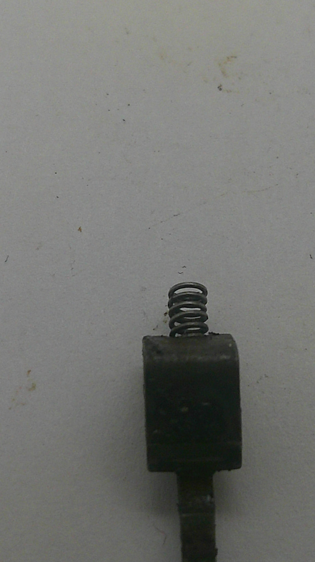A close-up of a small dark metal component with a coiled spring next to it, photographed on a plain off-white surface.
