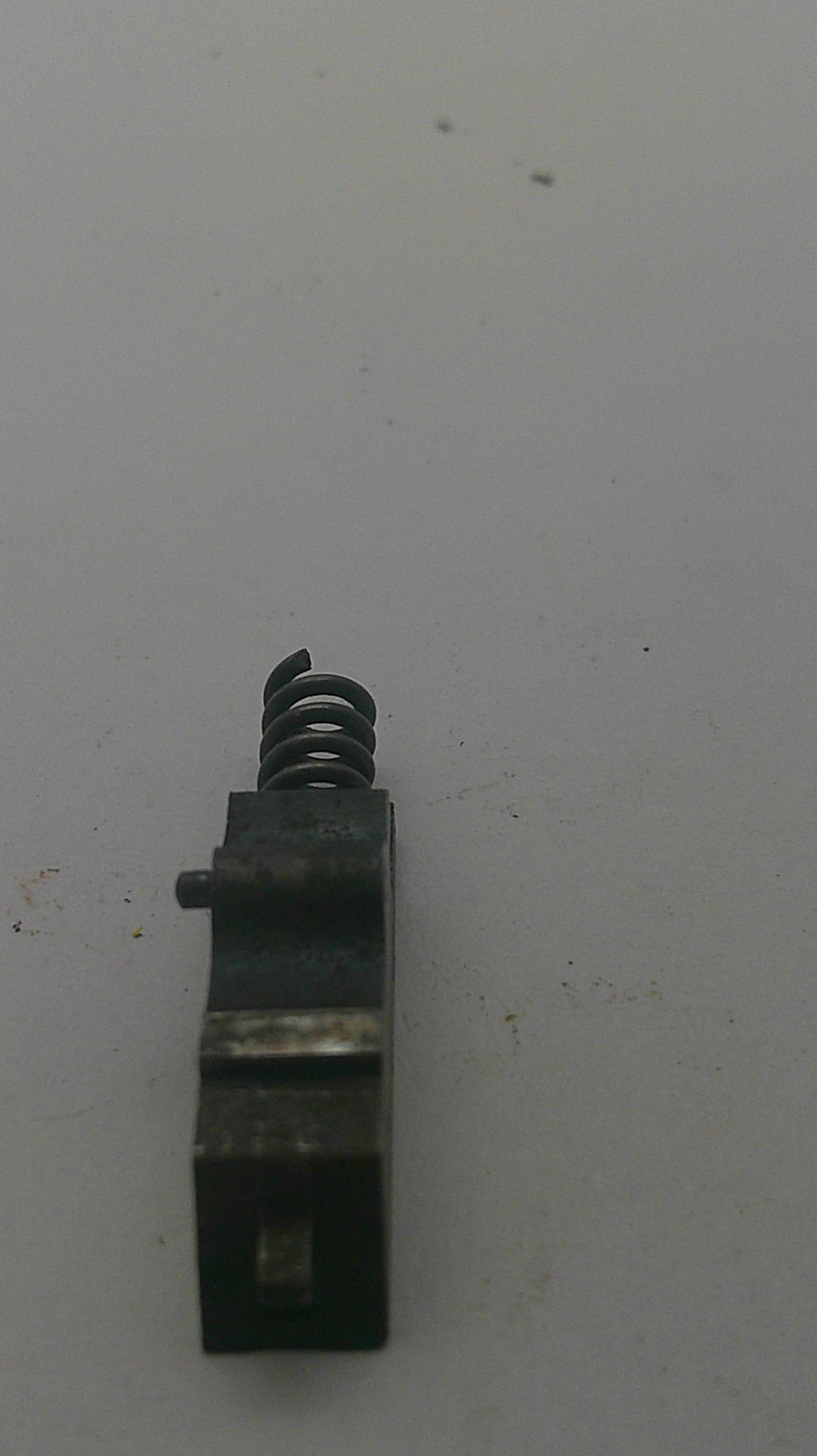 A small black metal mechanical component with a coiled spring, photographed on a plain off-white surface.